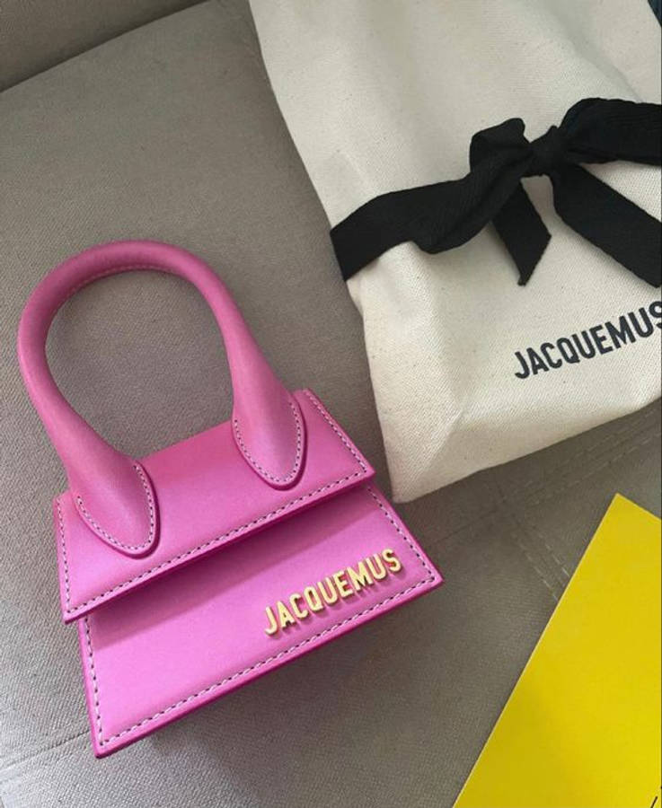 Handbag 桌布 壁紙 — 一個充滿活力的粉紅色Jacquemus手提包，帶有金色字母，旁邊是其品牌白色防塵袋，放在灰色布料表面上。