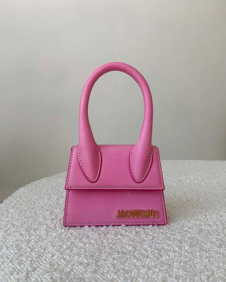 Handbag 桌布 壁紙 — 一個充滿活力的粉紅色Jacquemus手提包，帶有彎曲手柄，居中放置在帶紋理的白色表面上，背景為純色。