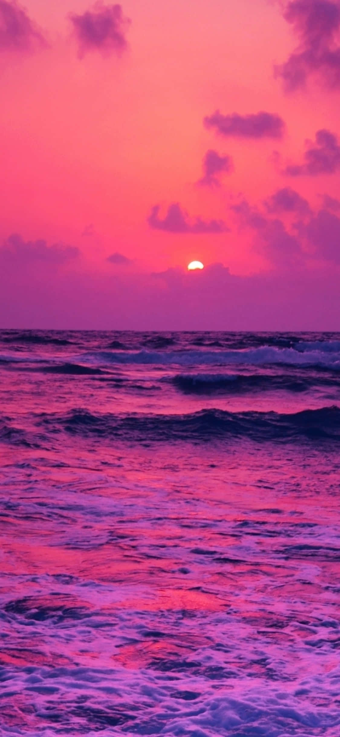 Pink Beach Sunset 桌布 壁紙 — 一個充滿活力的粉紅色和紫色日落，照耀著翻騰的海洋，低垂的太陽在海浪上投下火熱的色彩。