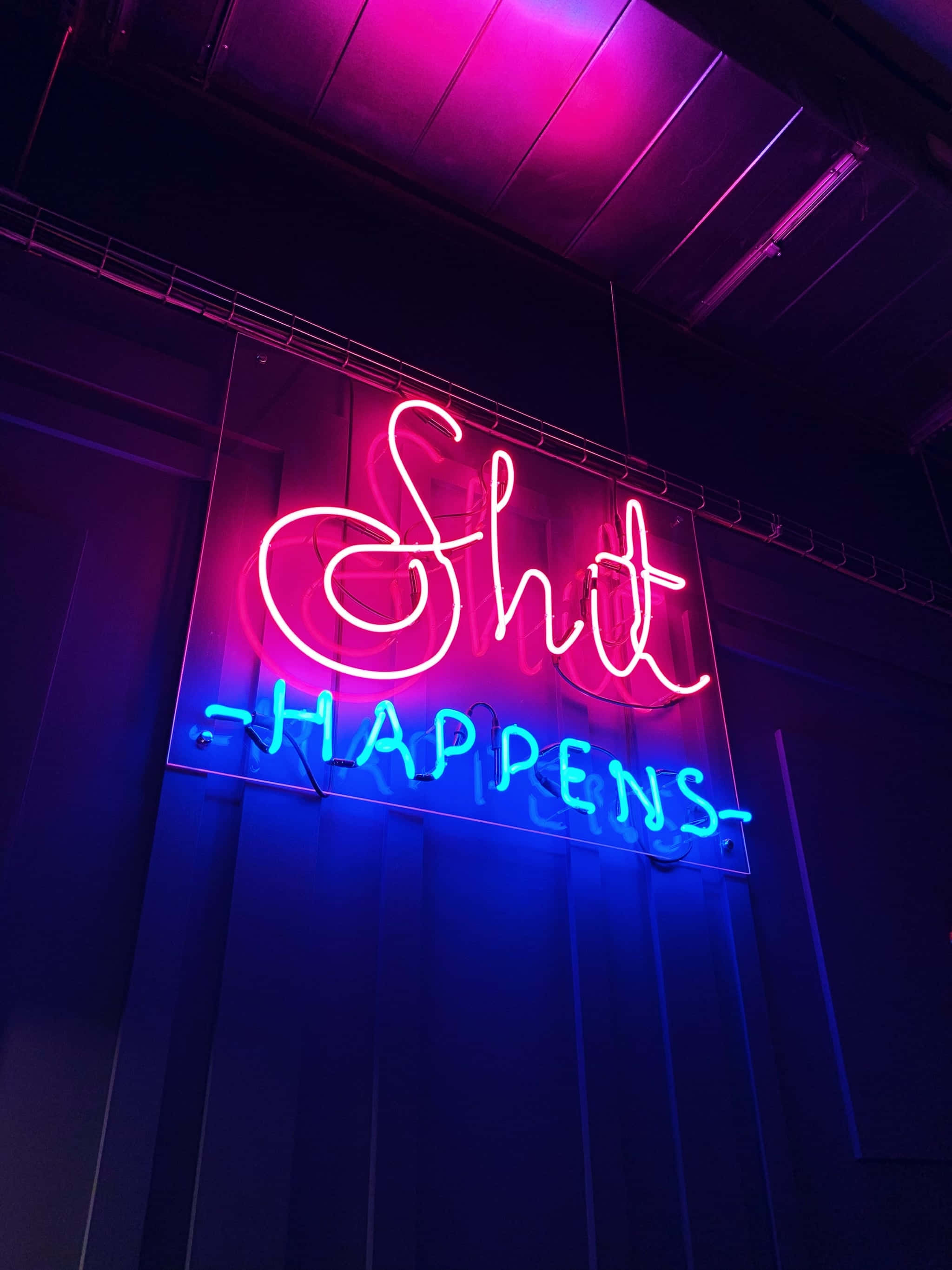 Neon Aesthetic Phone 桌布 壁紙 — 一個充滿活力的粉紅色和藍色霓虹燈牌，上面寫著「Shit Happens」，在黑暗的工業天花板上發光。