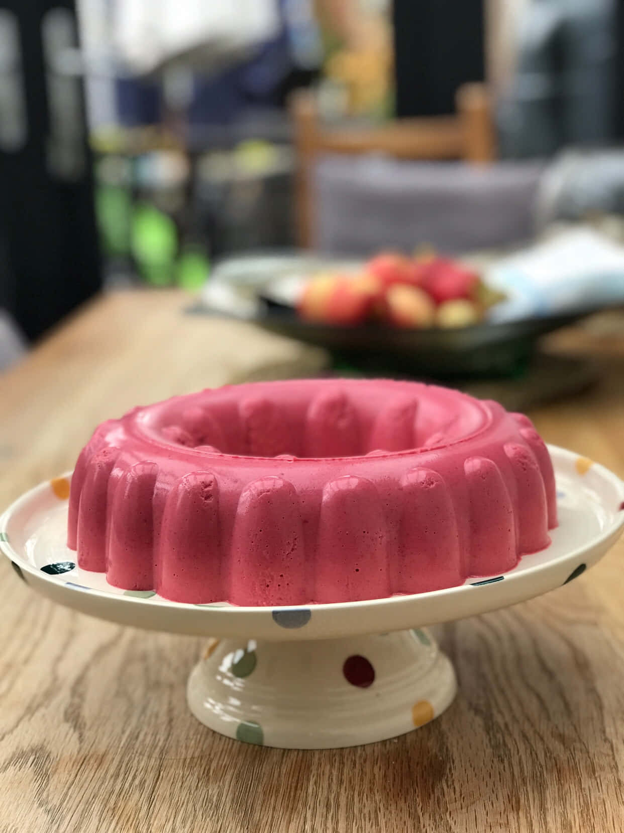 Blancmange 桌布 壁紙 — 一個充滿活力的粉紅色圓環蛋糕放在一個圓點蛋糕架上，置於木桌上，背景有模糊的水果。