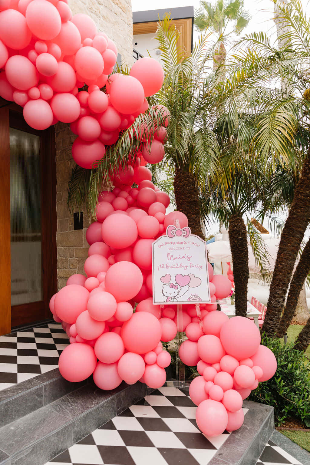 Hello Kitty Party 桌布 壁紙 — 一個充滿活力的粉紅色氣球拱門裝飾著格子圖案的庭院入口，旁邊有棕櫚樹，用於Hello Kitty生日派對。
