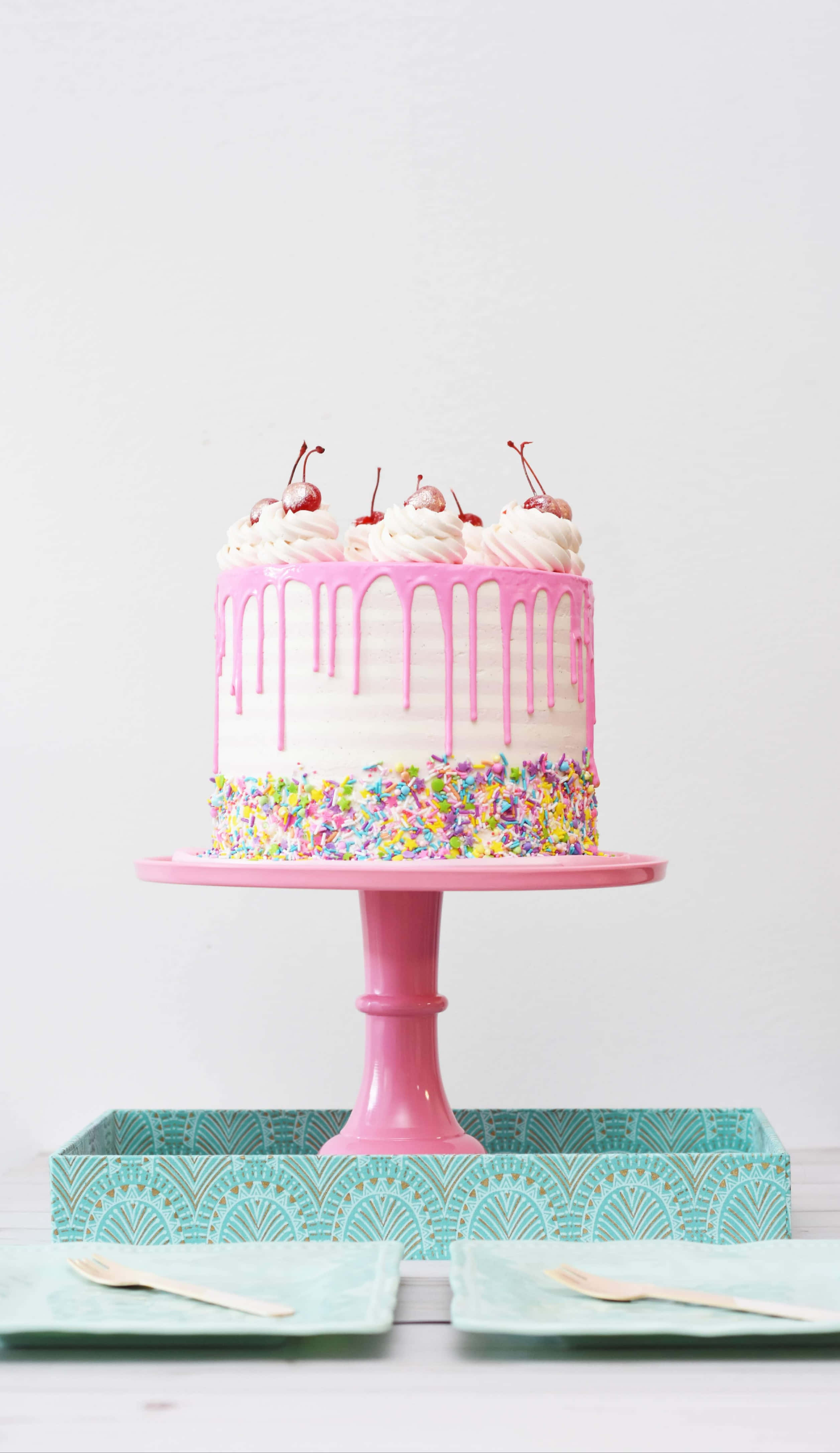 Cake Fondant 桌布 壁紙 — 一個充滿活力的粉紅色滴蛋糕，裝飾著色彩繽紛的糖粒和櫻桃，優雅地放在一個相配的架子上，背景是潔淨的白色。