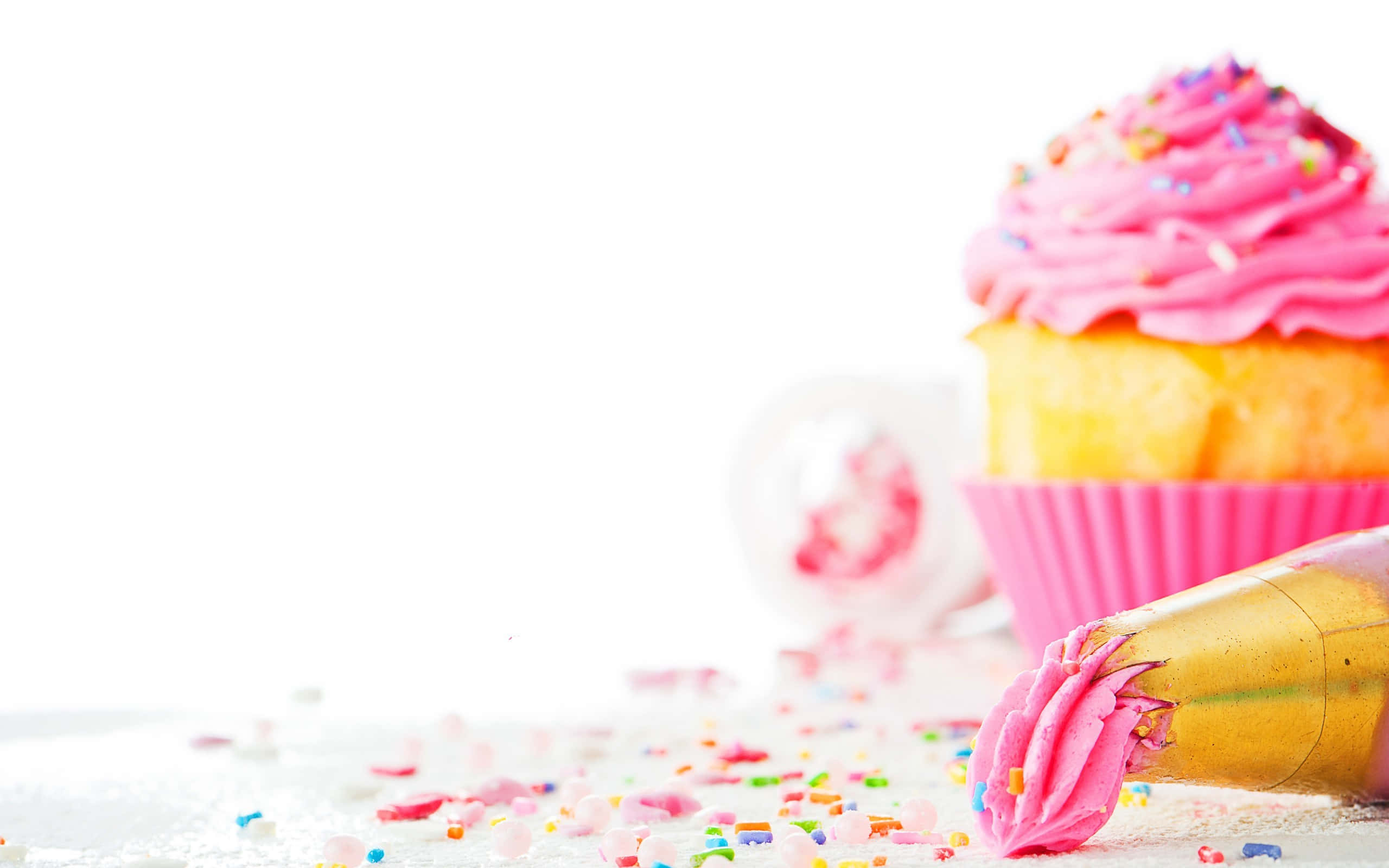Cute Cupcake 桌布 壁紙 — 一個充滿活力的粉紅色糖霜杯子蛋糕，上面有彩色糖粒，旁邊放著一個擠花袋，背景是明亮的白色。