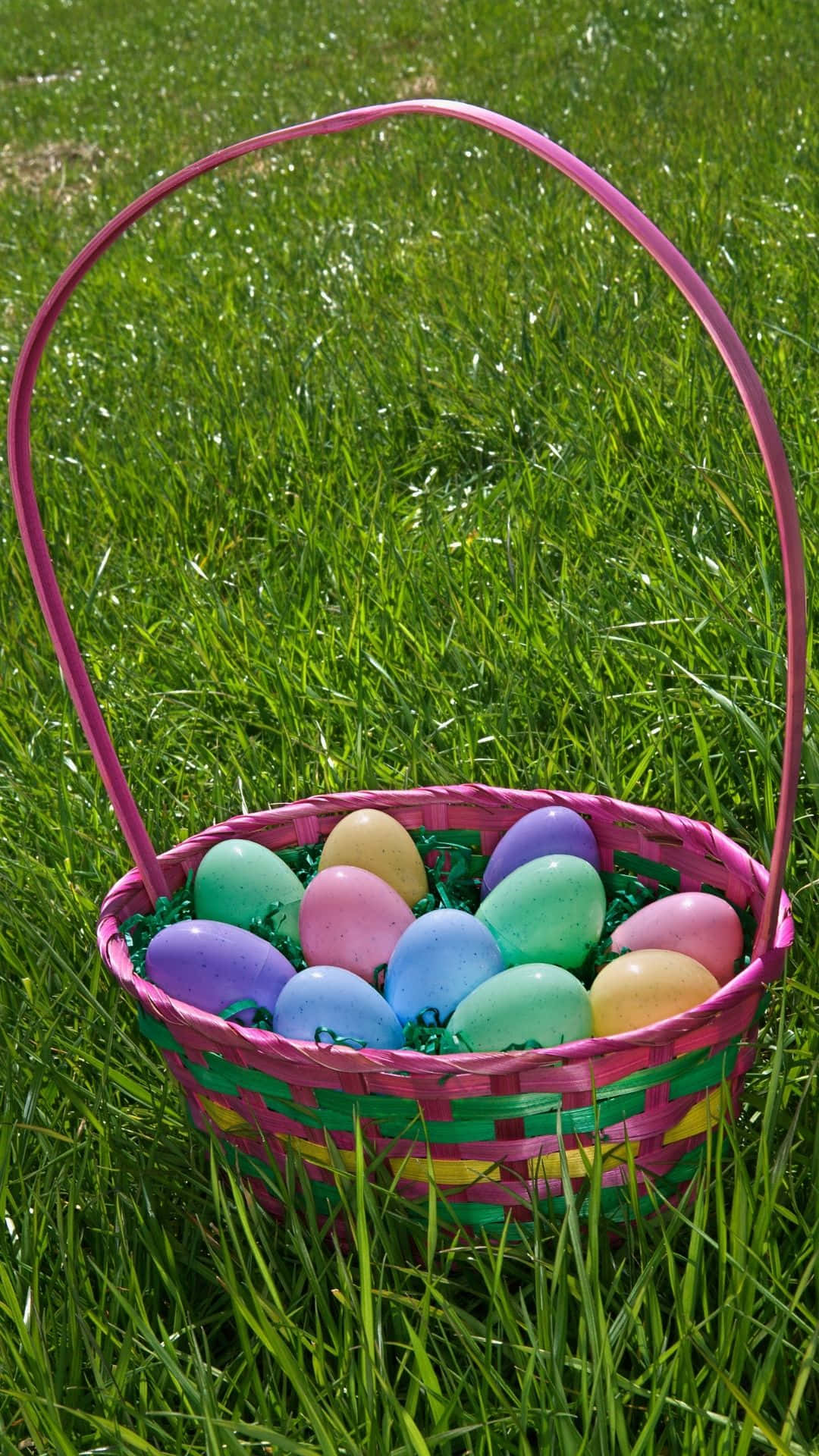 Easter Basket 桌布 壁紙 — 一個充滿活力的粉紅色編織籃子，裝滿了色彩繽紛的復活節彩蛋，在明亮的陽光下放在茂盛的綠草地上。