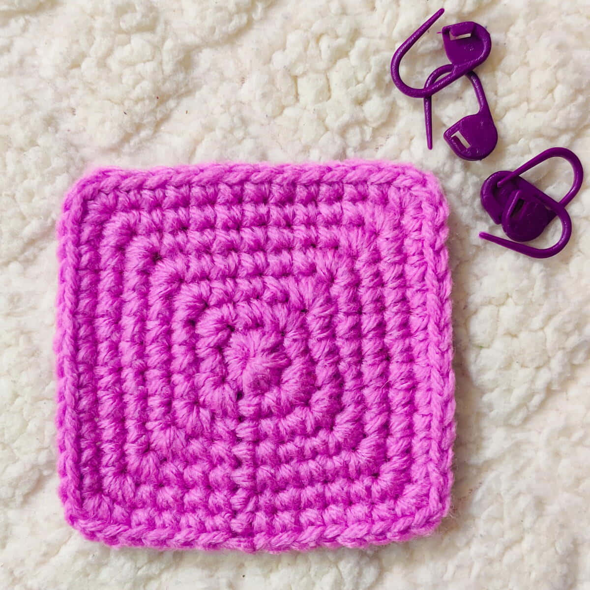 Crochet 桌布 壁紙 — 一個充滿活力的粉紅色鉤針編織正方形，帶有同心螺旋針跡，放在一個毛茸茸的白色表面上，旁邊有兩個紫色塑料針跡標記。
