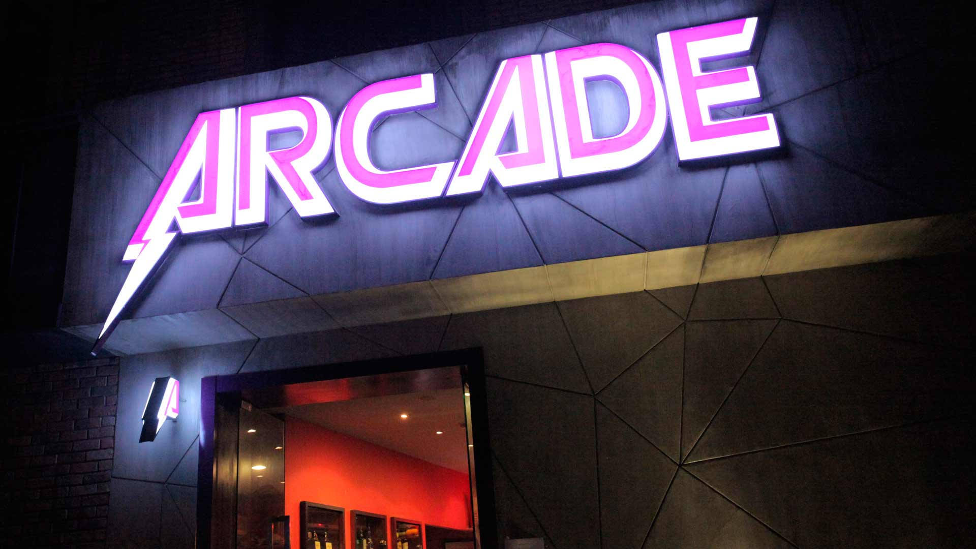 arcade 桌布 壁紙 — 一個充滿活力的粉紅色霓虹燈「ARCADE」標誌在夜間對著深色幾何外牆發光，照亮了復古遊戲場所的入口。