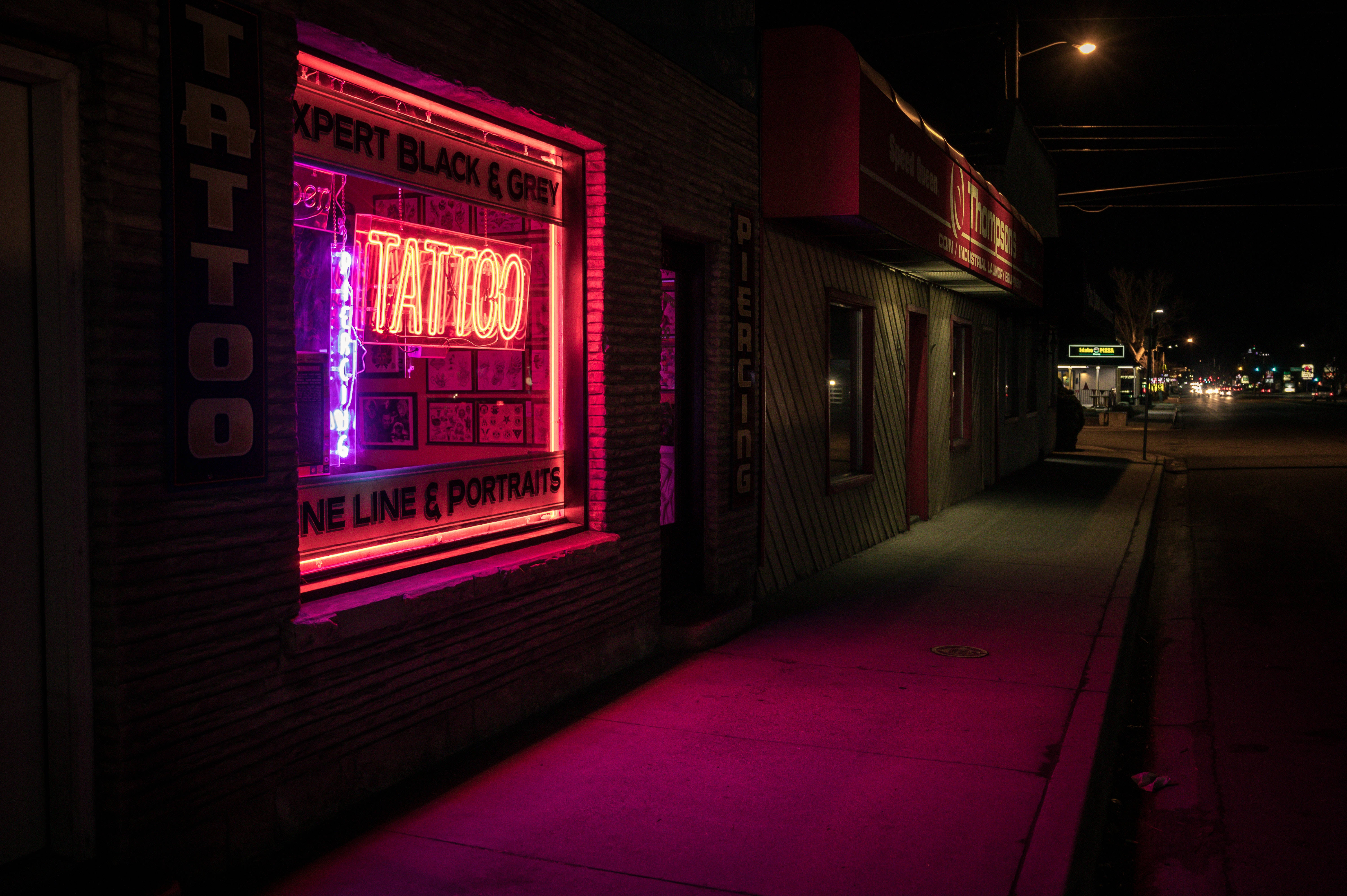 neon sign 桌布 壁紙 — 一個充滿活力的粉紅色霓虹紋身店招牌在漆黑的夜晚中發光，在人行道和磚牆上投射出超現實的光芒。