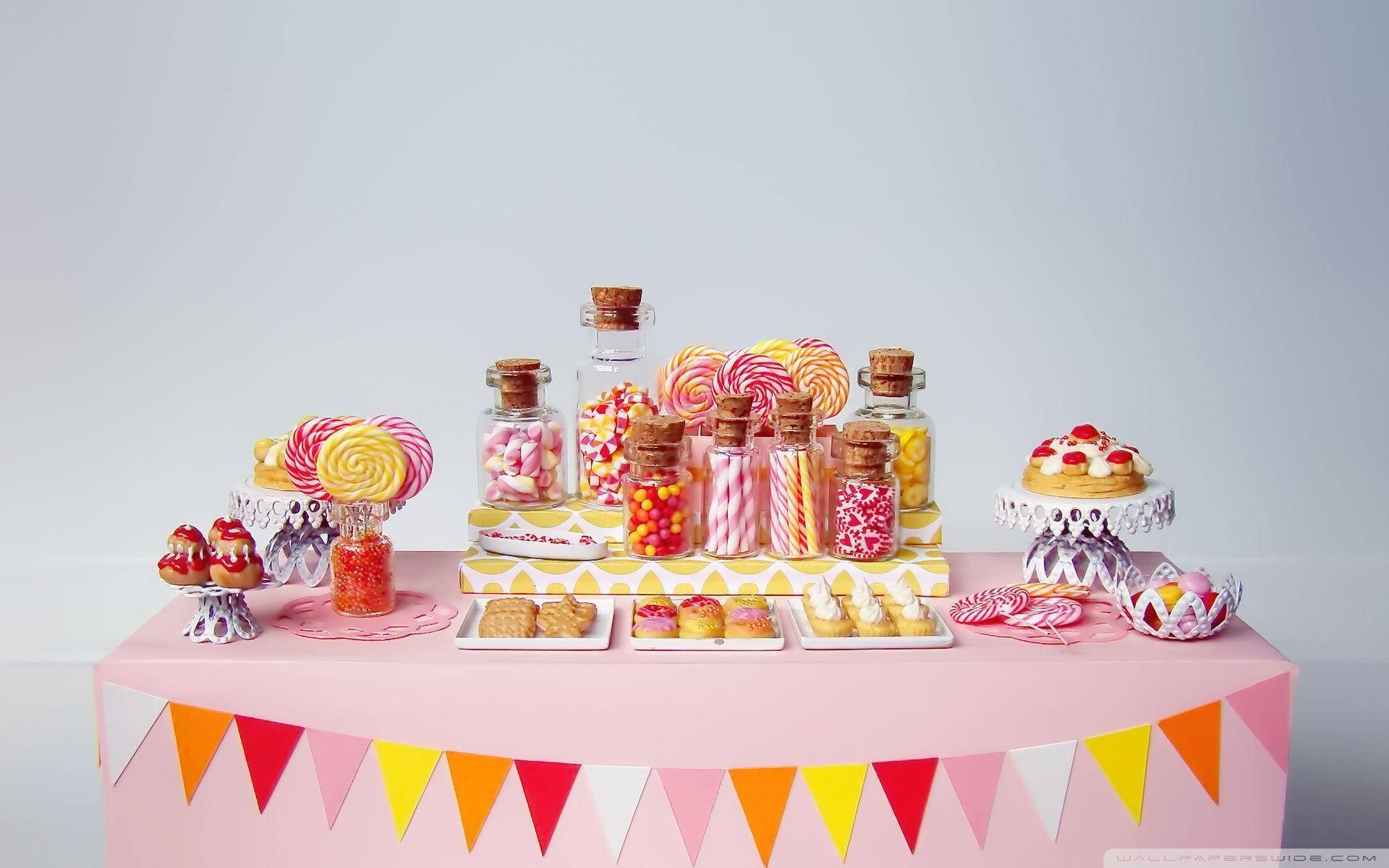 Desserts 桌布 壁紙 — 一個充滿活力的糖果自助餐，有棒棒糖、罐子、餅乾和粉彩色裝飾，放在粉紅色桌子上，背景為白色。