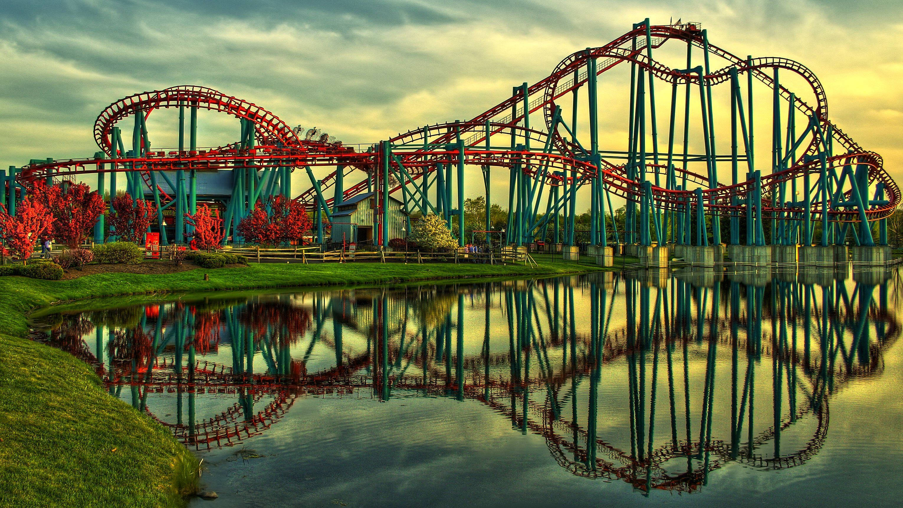 Roller Coaster 桌布 壁紙 — 一個充滿活力的紅色和藍綠色雲霄飛車完美地倒映在平靜的池塘中，正值黃金時段，附近有秋天的樹葉。