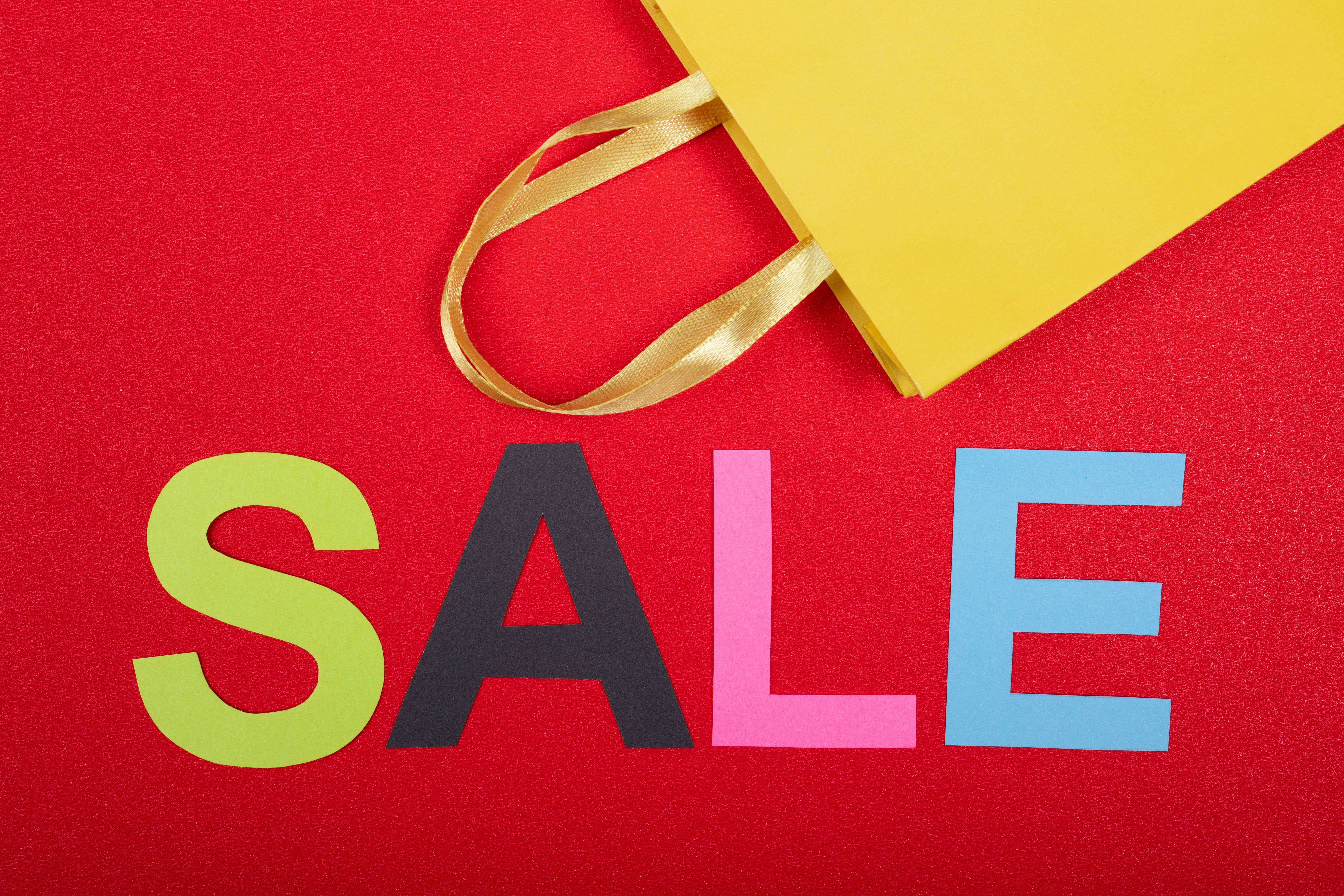 Sales 桌布 壁紙 — 一個充滿活力的紅色紋理表面上，用彩色塊狀字母寫著「SALE」，附近有一個黃色購物袋和金色絲帶。