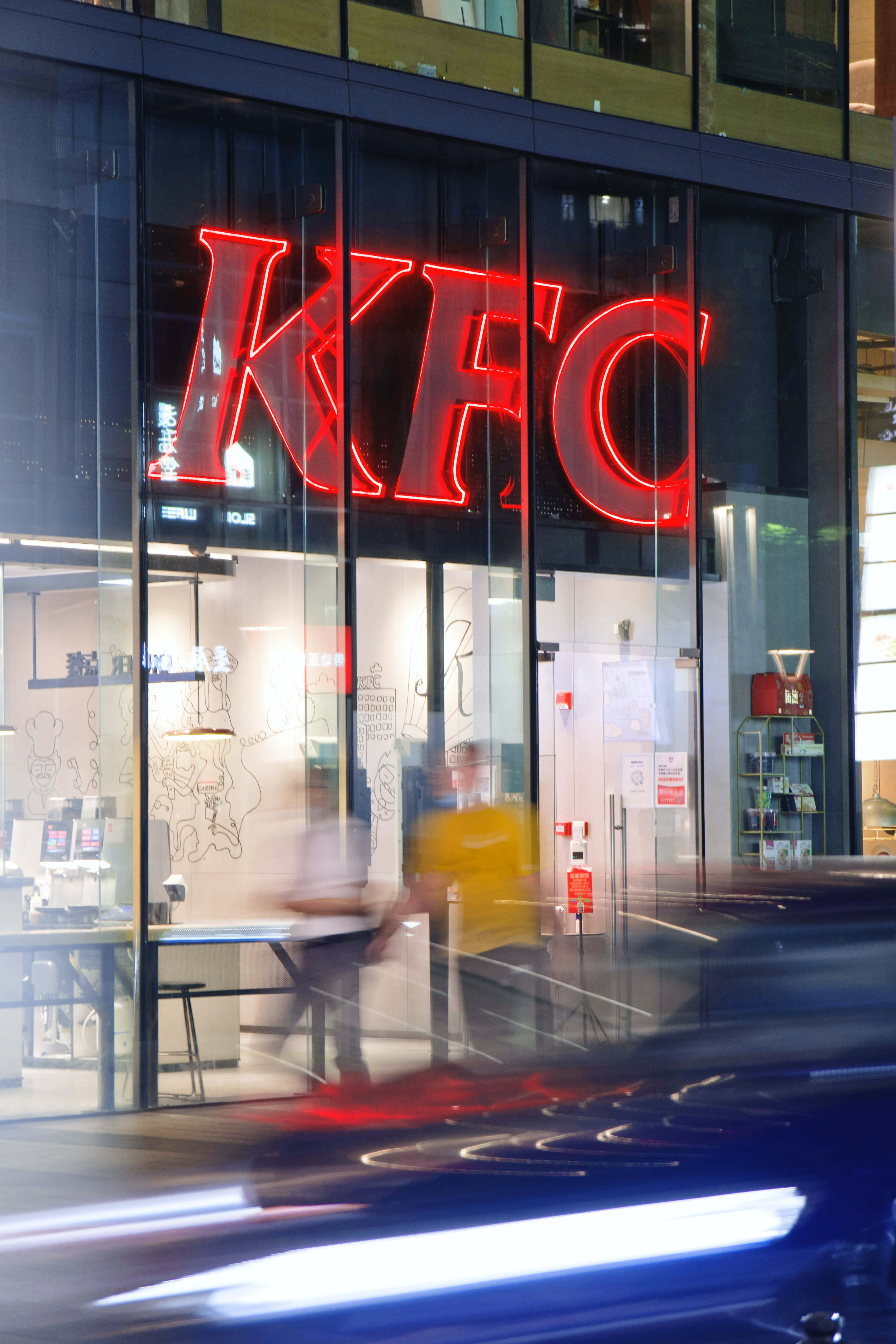 Kfc 桌布 壁紙 — 一個充滿活力的紅色霓虹燈KFC標誌透過玻璃店面發光，模糊的行人和一輛高速行駛的汽車在夜晚疾馳而過。