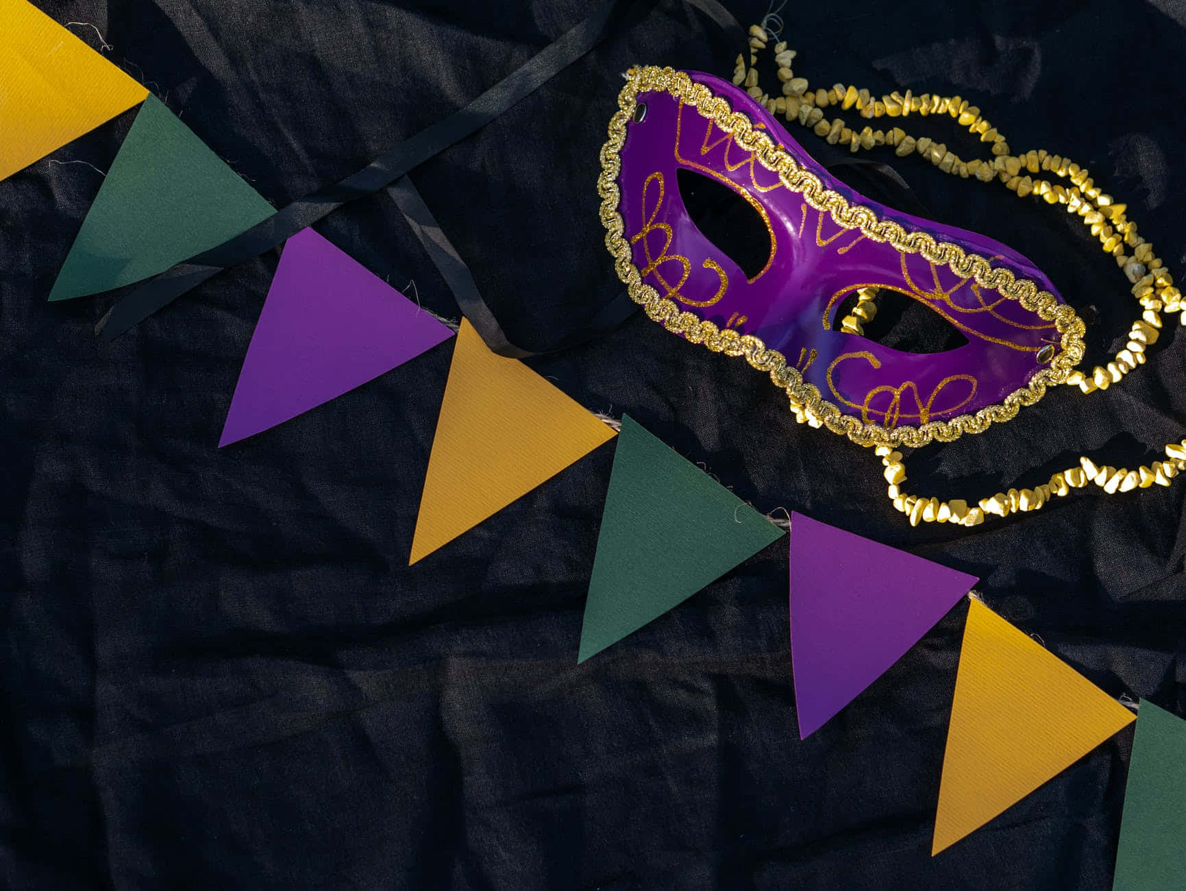Mardi Gras 桌布 壁紙 — 一個充滿活力的紫色、金色和綠色狂歡節面具，旁邊是節日三角旗，放在深色布料背景上。
