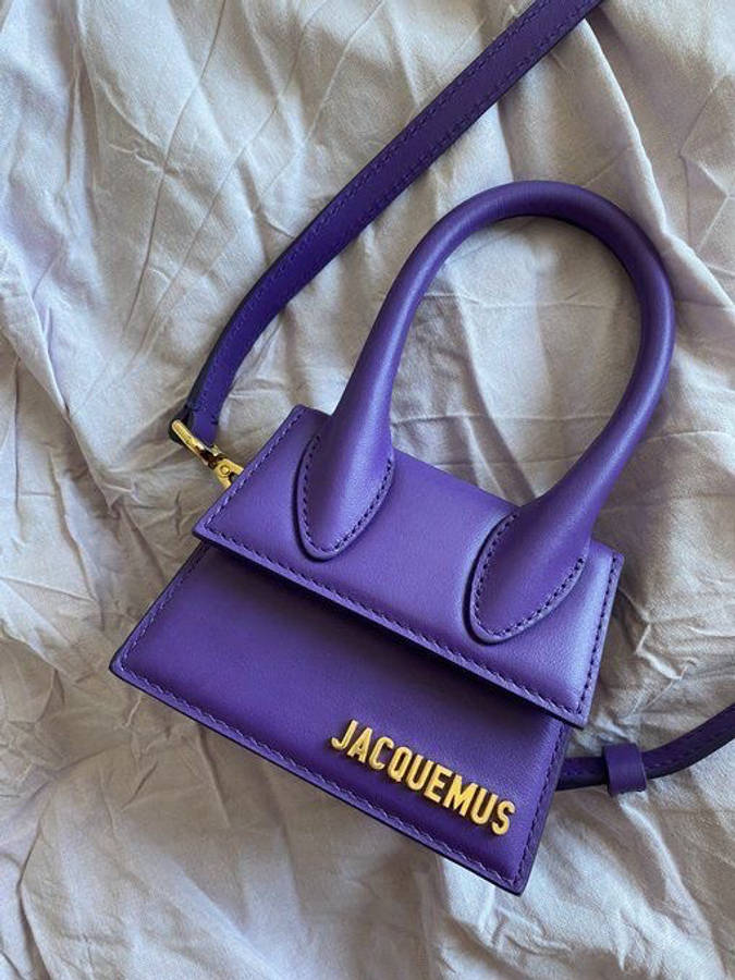 Handbag 桌布 壁紙 — 一個充滿活力的紫色Jacquemus手提包，帶有金色字母，放在皺巴巴的米白色布料上。