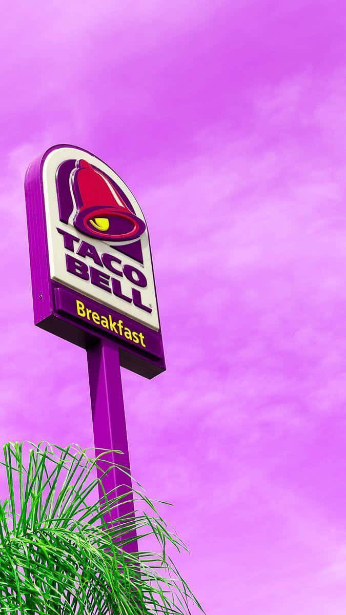 塔可鐘 桌布 壁紙 — 一個充滿活力的紫色Taco Bell招牌，上面有「Breakfast」字樣，在超現實的粉紅色天空和底部綠色棕櫚葉的襯托下高高聳立。