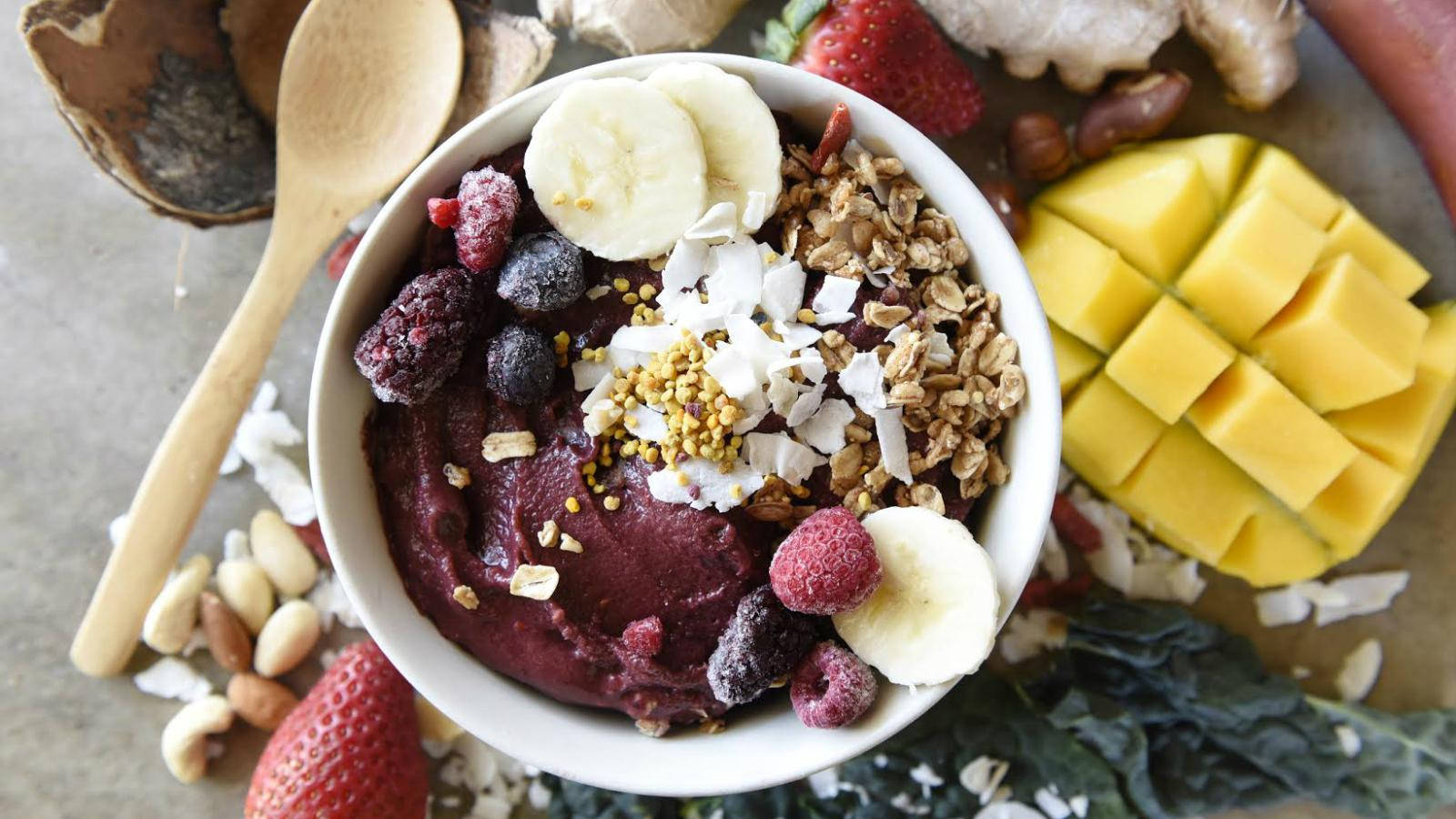 Healthy Food 桌布 壁紙 — 一個充滿活力的紫色冰沙碗，上面鋪滿香蕉片、燕麥片、椰子片和新鮮莓果，旁邊有切塊芒果、草莓和堅果，放在一個鄉村風托盤上。