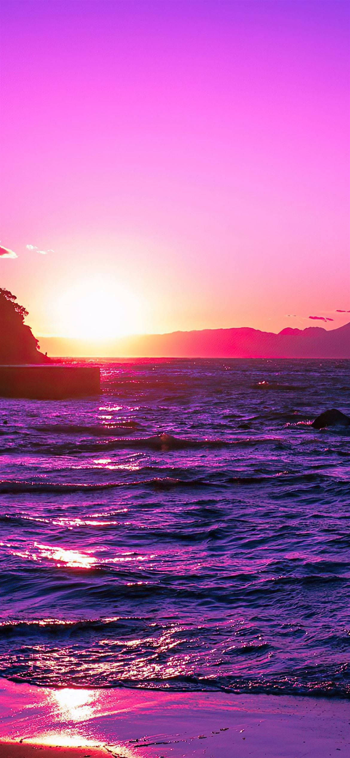 Pink Sunset iPhone 桌布 壁紙 — 一個充滿活力的紫色和粉紅色日落，映照在波光粼粼的海洋上，遠方有山脈，地平線閃耀著光芒。