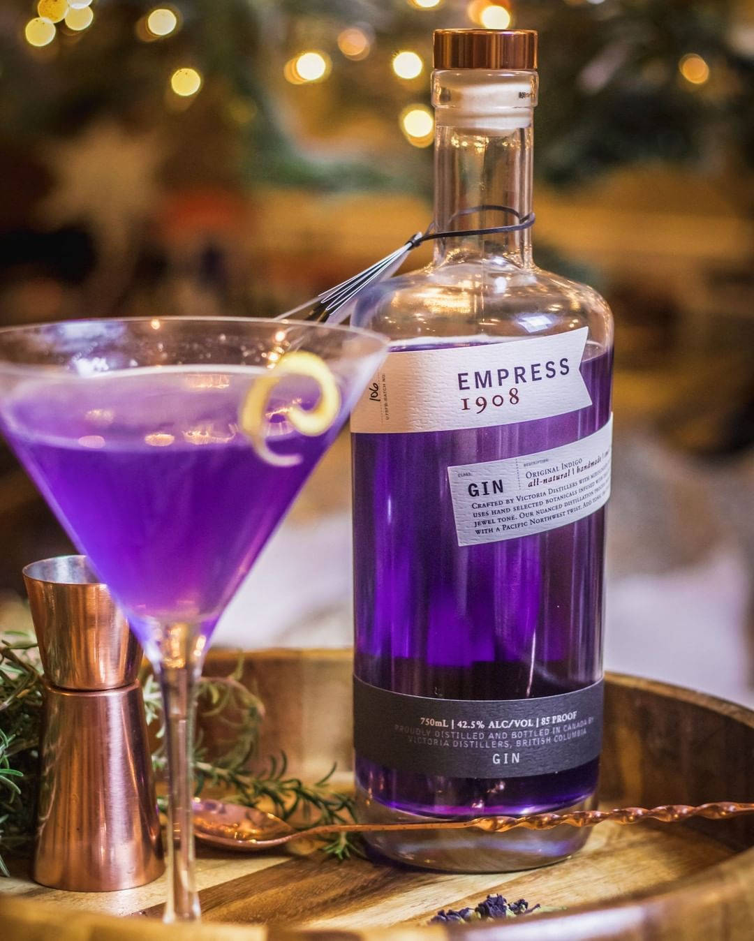 alcohol 桌布 壁紙 — 一個充滿活力的紫色琴酒瓶和以檸檬扭紋裝飾的雞尾酒放在木托盤上，背景有銅製酒吧工具和節日燈飾。