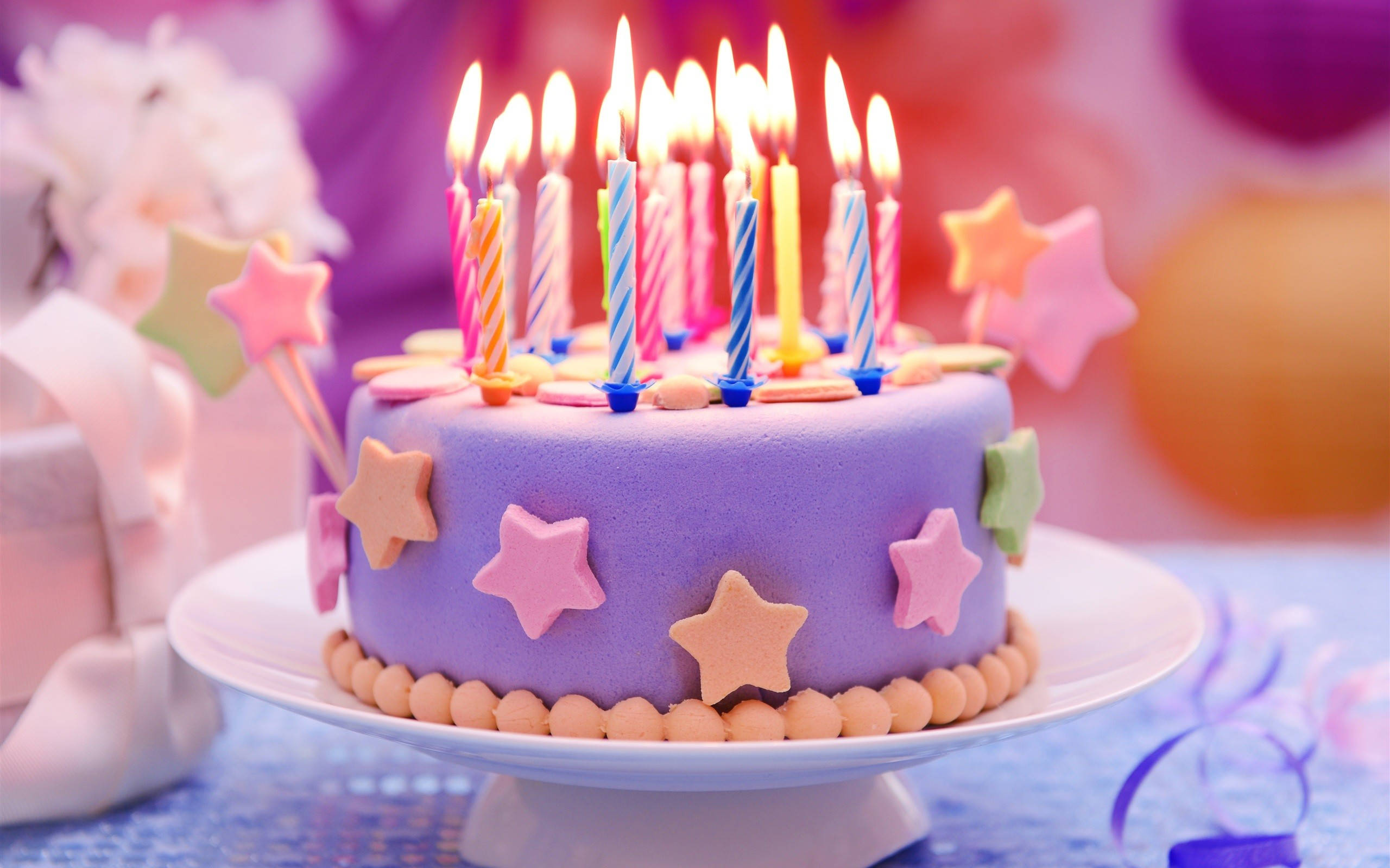 Cake 桌布 壁紙 — 一個充滿活力的紫色生日蛋糕，裝飾著點燃的蠟燭和色彩繽紛的星星裝飾，放在一個基座上，背景是節日模糊的。