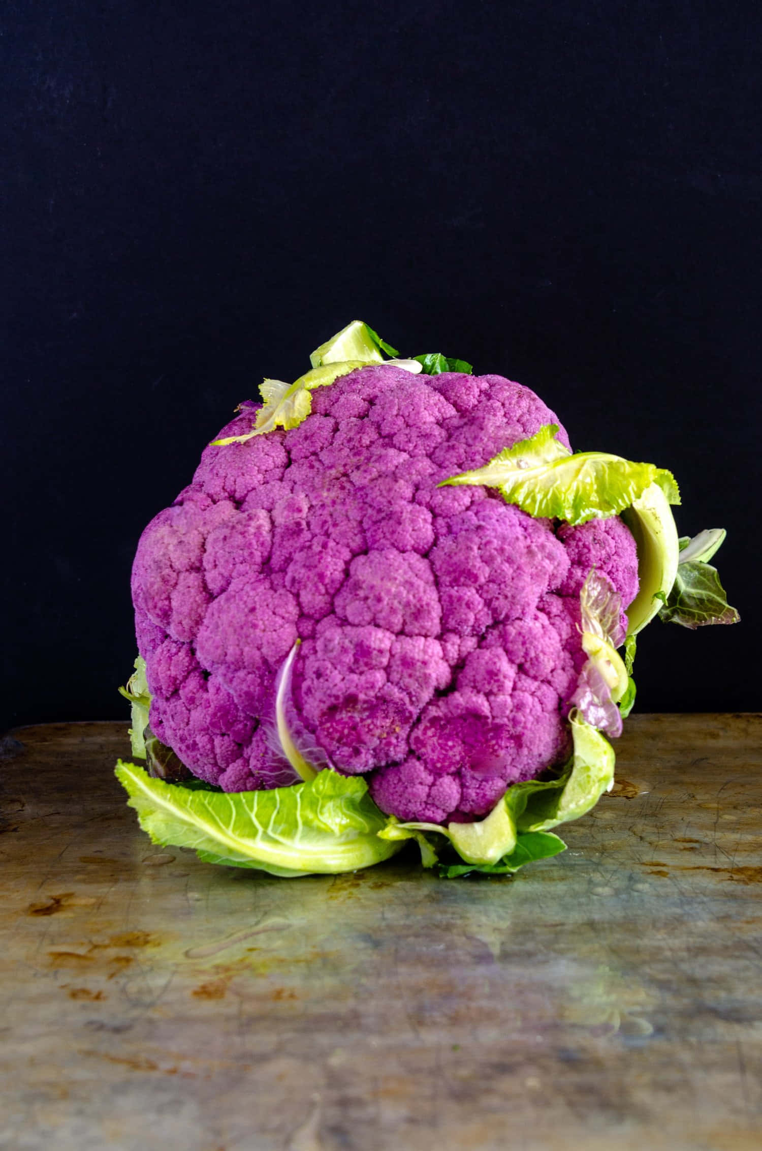 Purple Cauliflower 桌布 壁紙 — 一個充滿活力的紫色花椰菜，帶有新鮮的綠色葉子，放在一個風化的金屬表面上，背景是深色的。