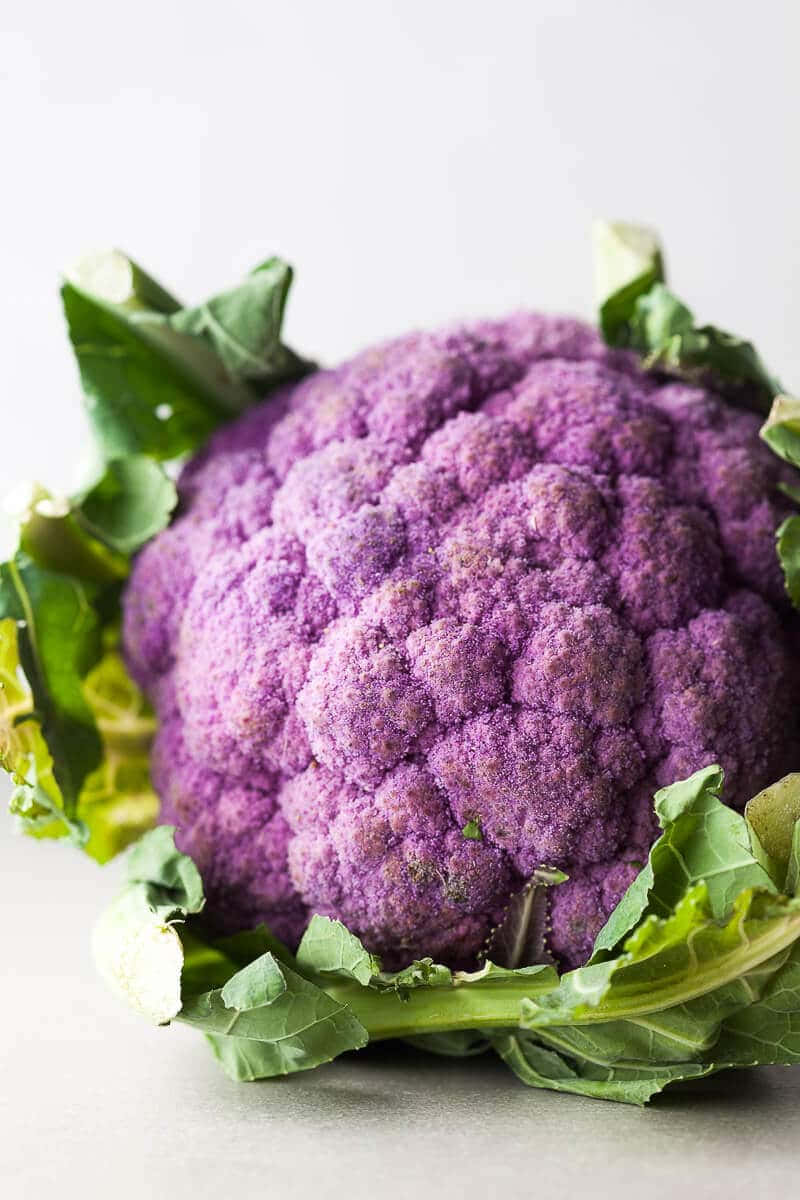 Purple Cauliflower 桌布 壁紙 — 一個充滿活力的紫色花椰菜，帶有新鮮的綠色葉子，在柔和的工作室燈光下放置在灰色中性表面上。