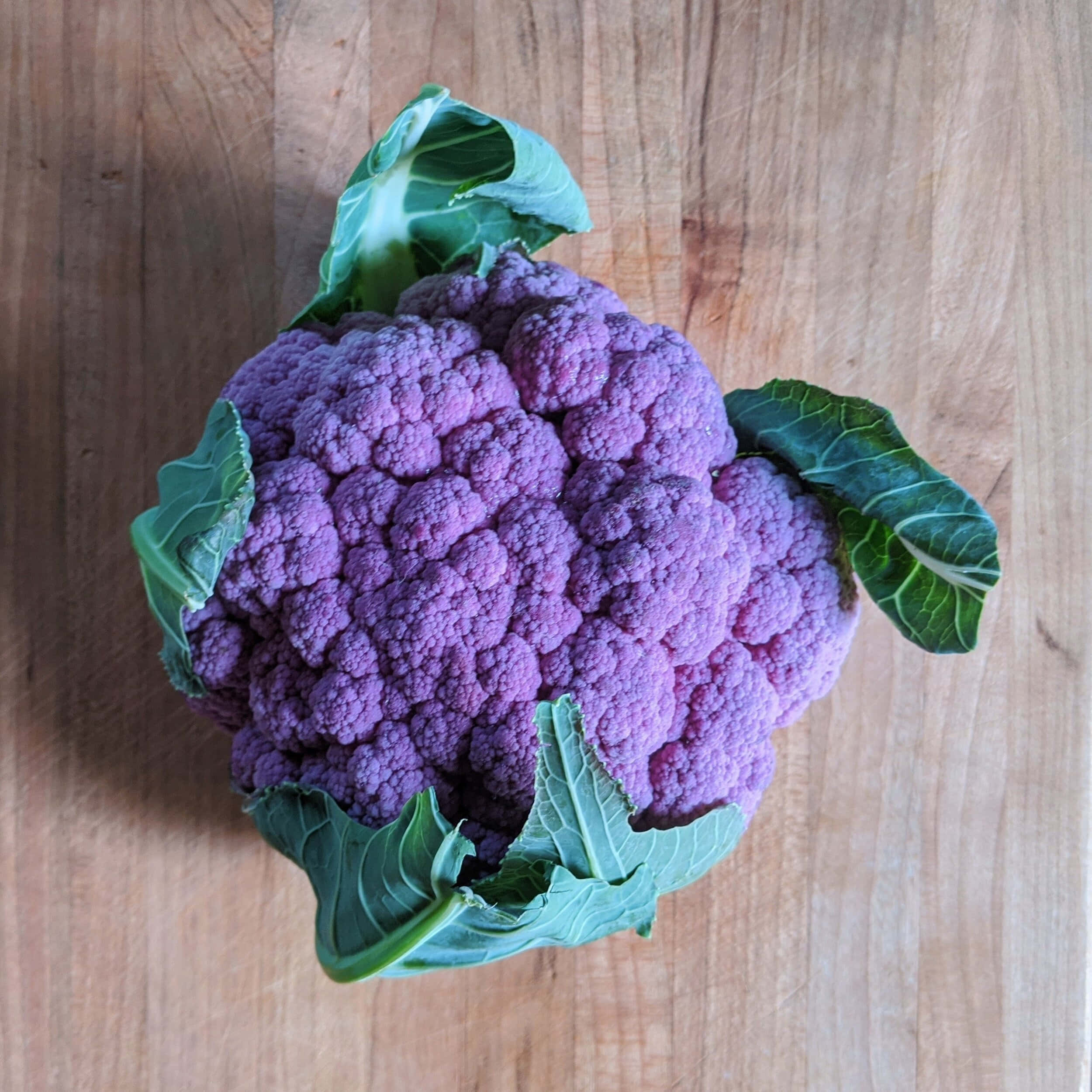 Purple Cauliflower 桌布 壁紙 — 一個充滿活力的紫色花椰菜，帶有綠色葉子，放在溫暖的木砧板上。