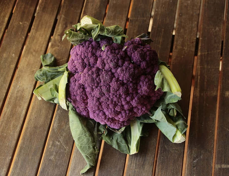 Purple Cauliflower 桌布 壁紙 — 一個充滿活力的紫色花椰菜，帶有綠色葉子，放在風化的木條表面上。