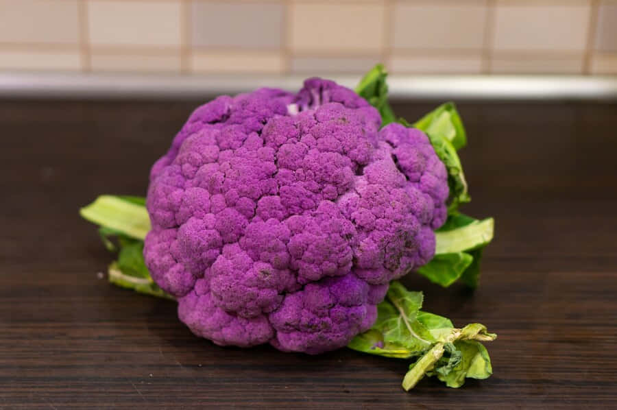 Purple Cauliflower 桌布 壁紙 — 一個充滿活力的紫色花椰菜，帶有綠色葉子，放在深色木質檯面上，背景是瓷磚牆。