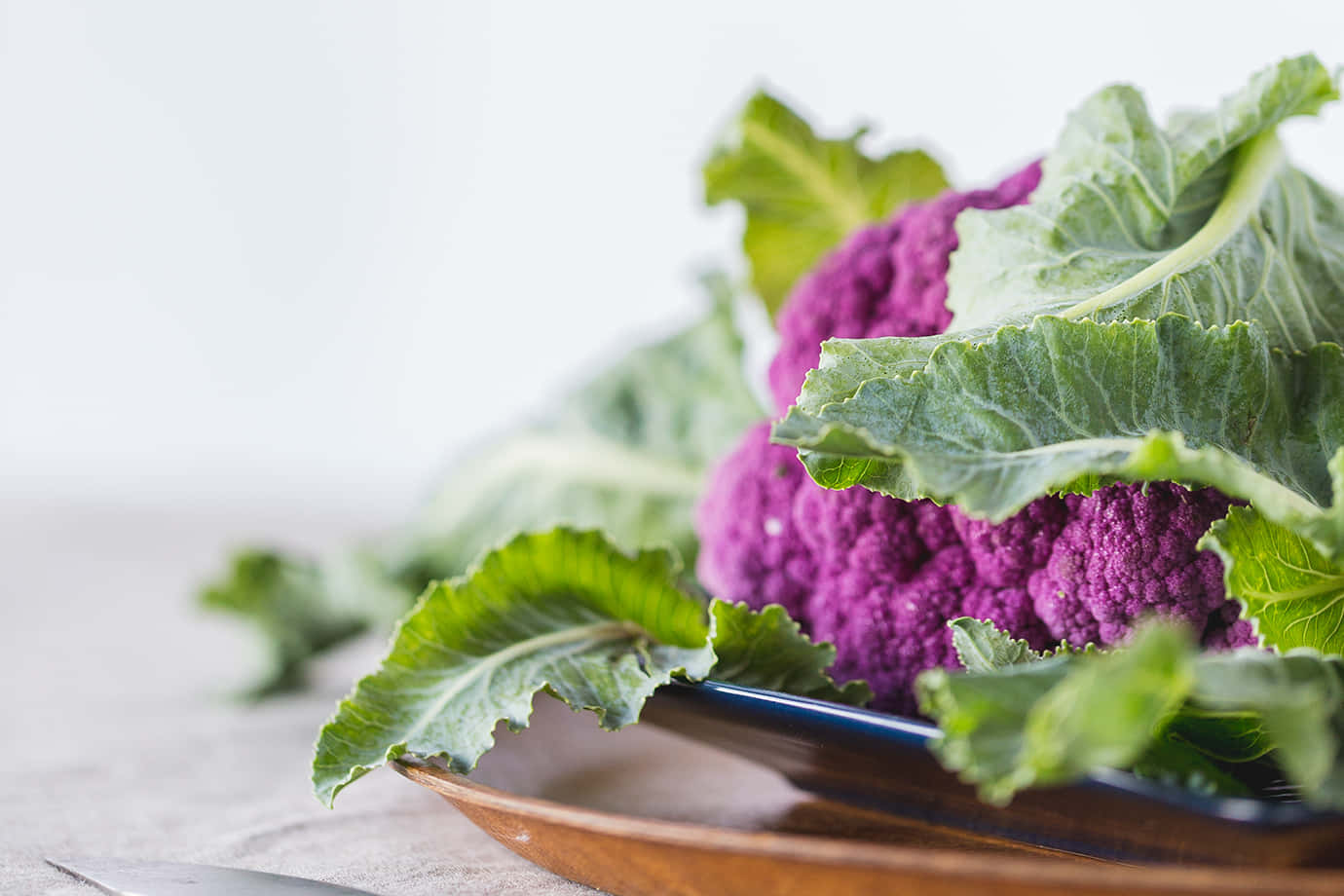 Purple Cauliflower 桌布 壁紙 — 一個充滿活力的紫色花椰菜，帶有茂盛的綠葉，放在木盤上，襯托著柔和的白色背景。