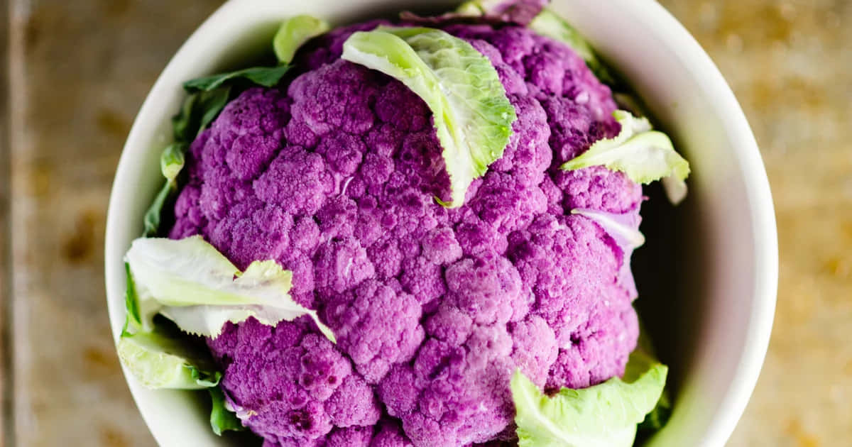 Purple Cauliflower 桌布 壁紙 — 一個充滿活力的紫色花椰菜放在白色碗中，搭配綠色葉子，從正上方近距離拍攝。