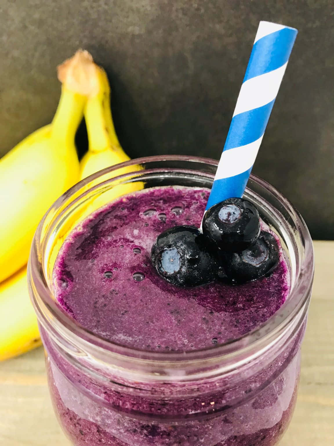 Blueberry Smoothie 桌布 壁紙 — 一個充滿活力的紫色藍莓香蕉冰沙，裝在玻璃罐中，上面放上新鮮藍莓和一條藍白條紋吸管。
