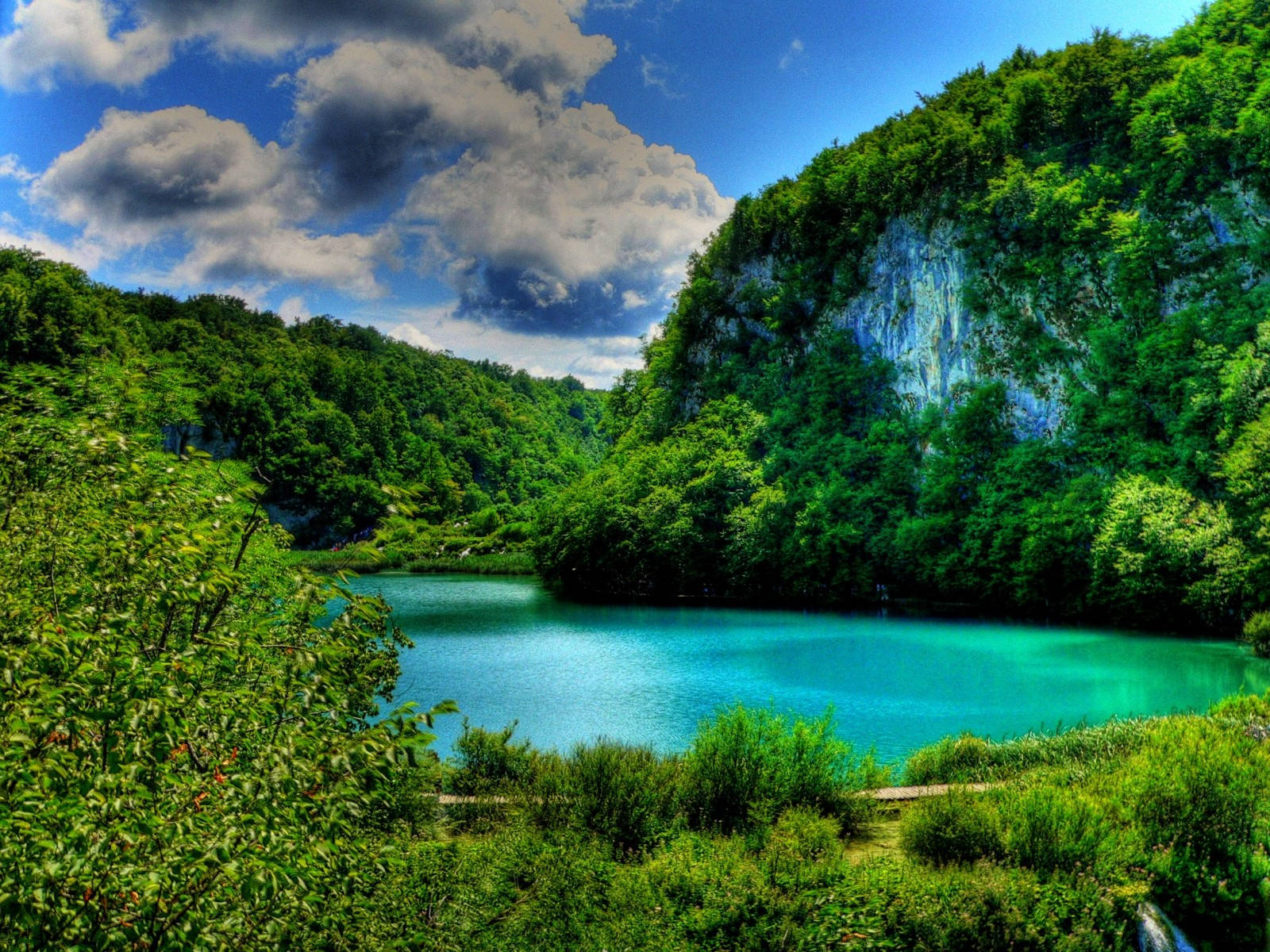 Croatia 桌布 壁紙 — 一個充滿活力的綠松石色湖泊， nestled 在茂密的綠色森林中，上方是戲劇性的藍天和蓬鬆的白雲。