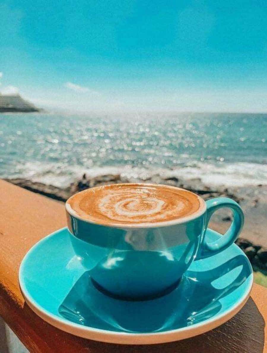 Morning Coffee 桌布 壁紙 — 一個充滿活力的綠松色咖啡杯，帶有拉花藝術，放在木欄杆上，俯瞰著清澈藍天下閃閃發光的大海。