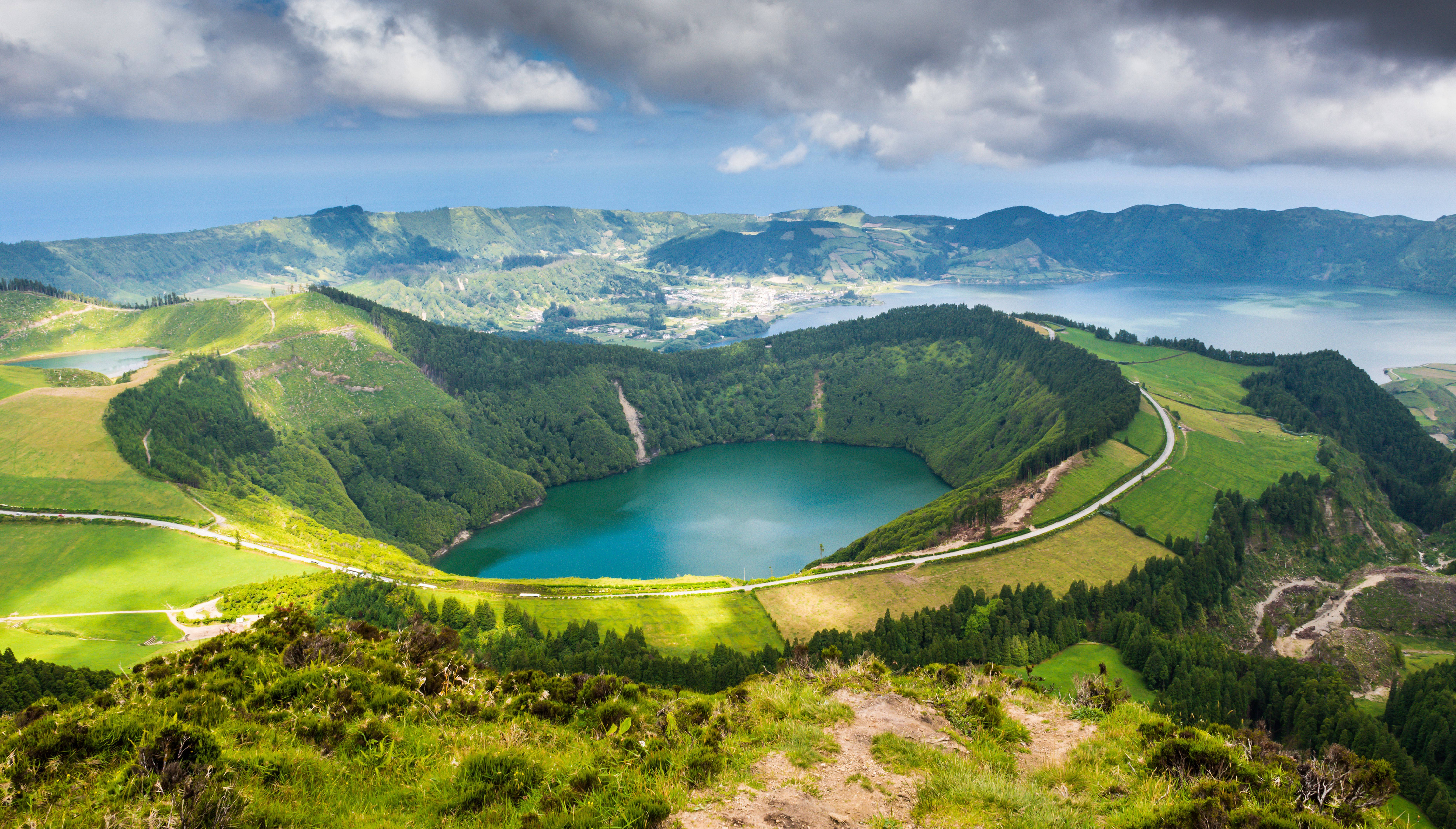 Portugal 桌布 壁紙 — 一個充滿活力的綠松色火山口湖，坐落在鬱鬱蔥蔥的綠色山丘中，上方是戲劇性的多雲天空。