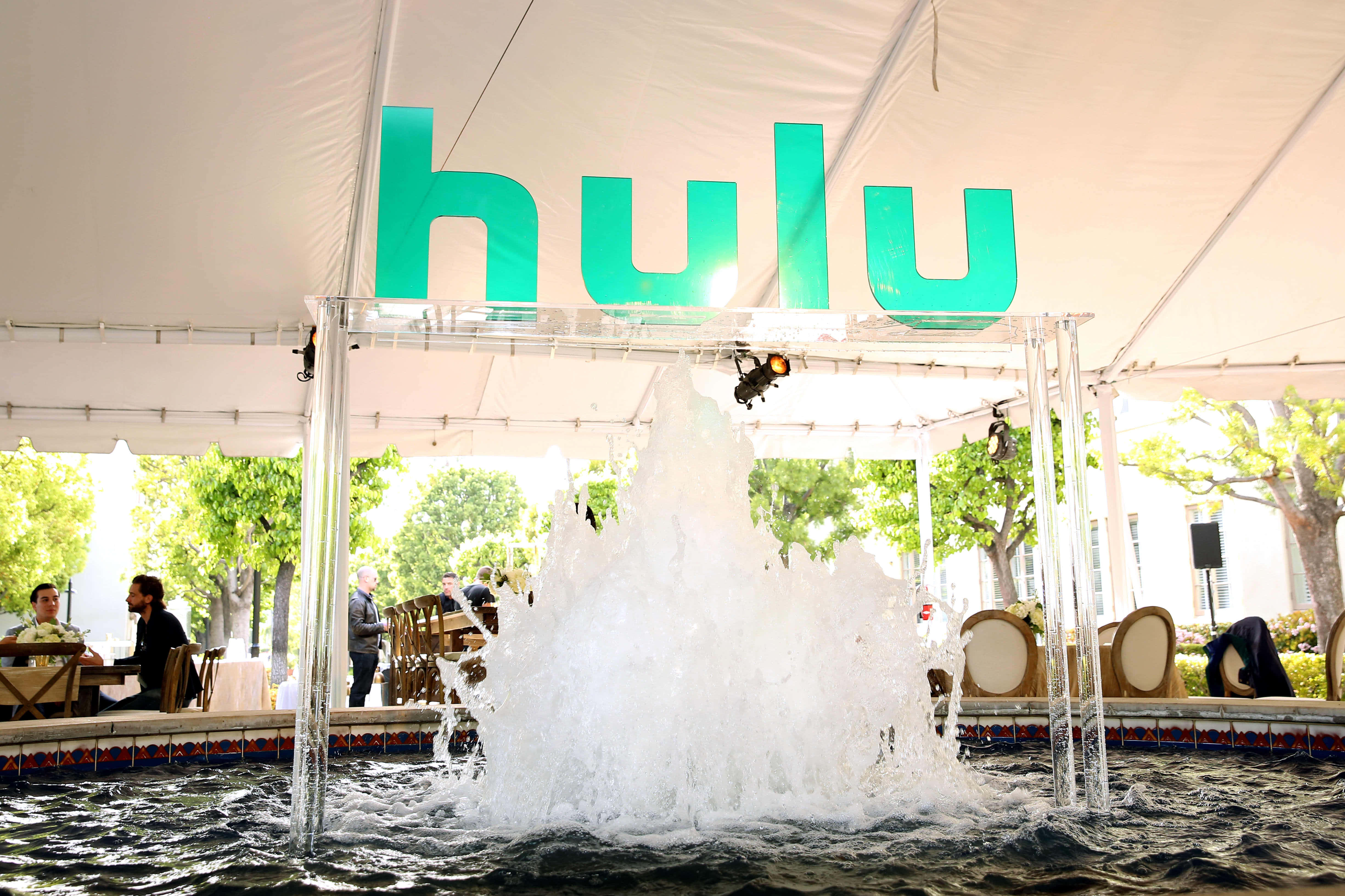 Hulu 桌布 壁紙 — 一個充滿活力的綠色Hulu標誌拱起在一個熱鬧的噴泉上方，下方是白色帳篷，附近有正在用餐的人們。