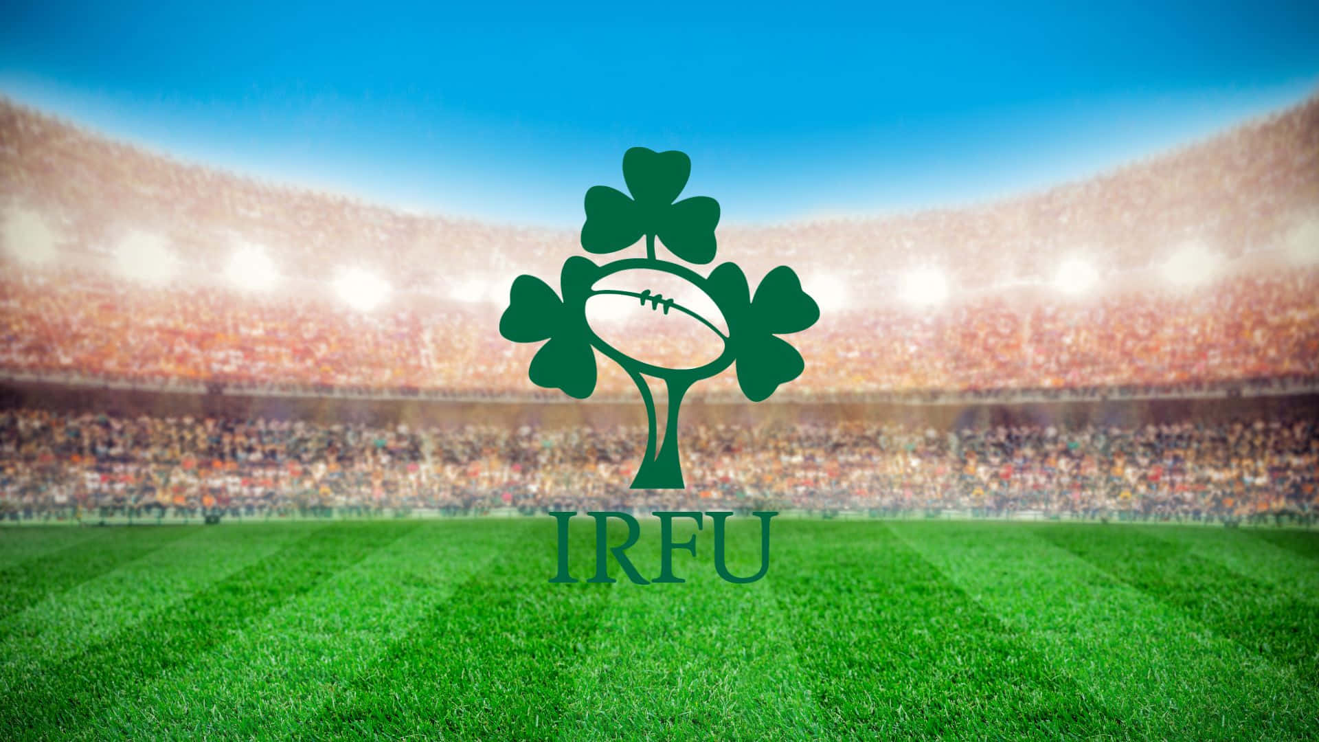 Ireland Rugby 桌布 壁紙 — 一個充滿活力的綠色三葉草標誌，中心有一個橄欖球，矗立在茂密的草地上，前方是座無虛席的體育場，天空湛藍清澈。