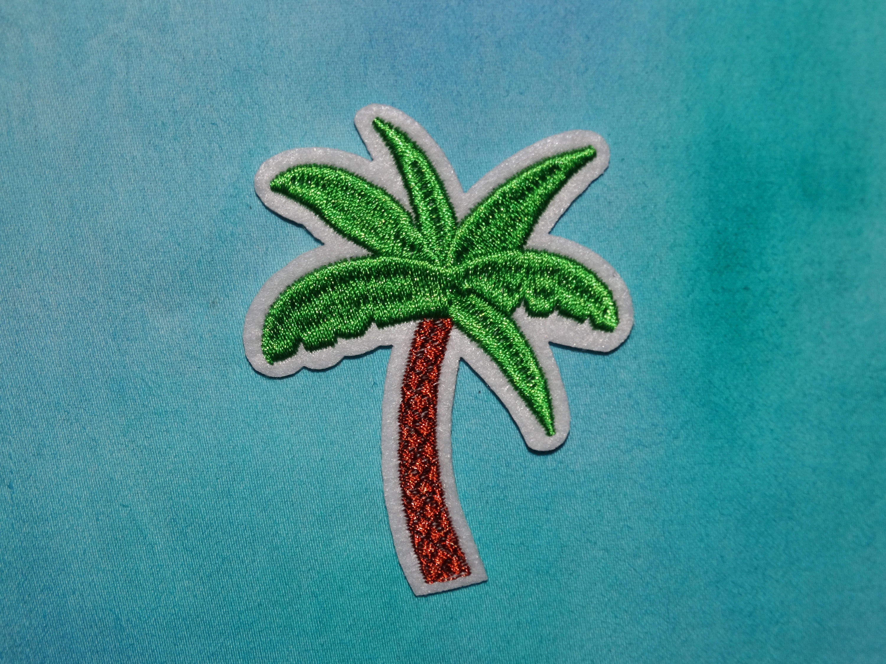 青色GIF 桌布 壁紙 — 一個充滿活力的綠色刺繡棕櫚樹補丁，帶有閃閃發光的紅色樹幹，位於有紋理的青綠色布料背景上。