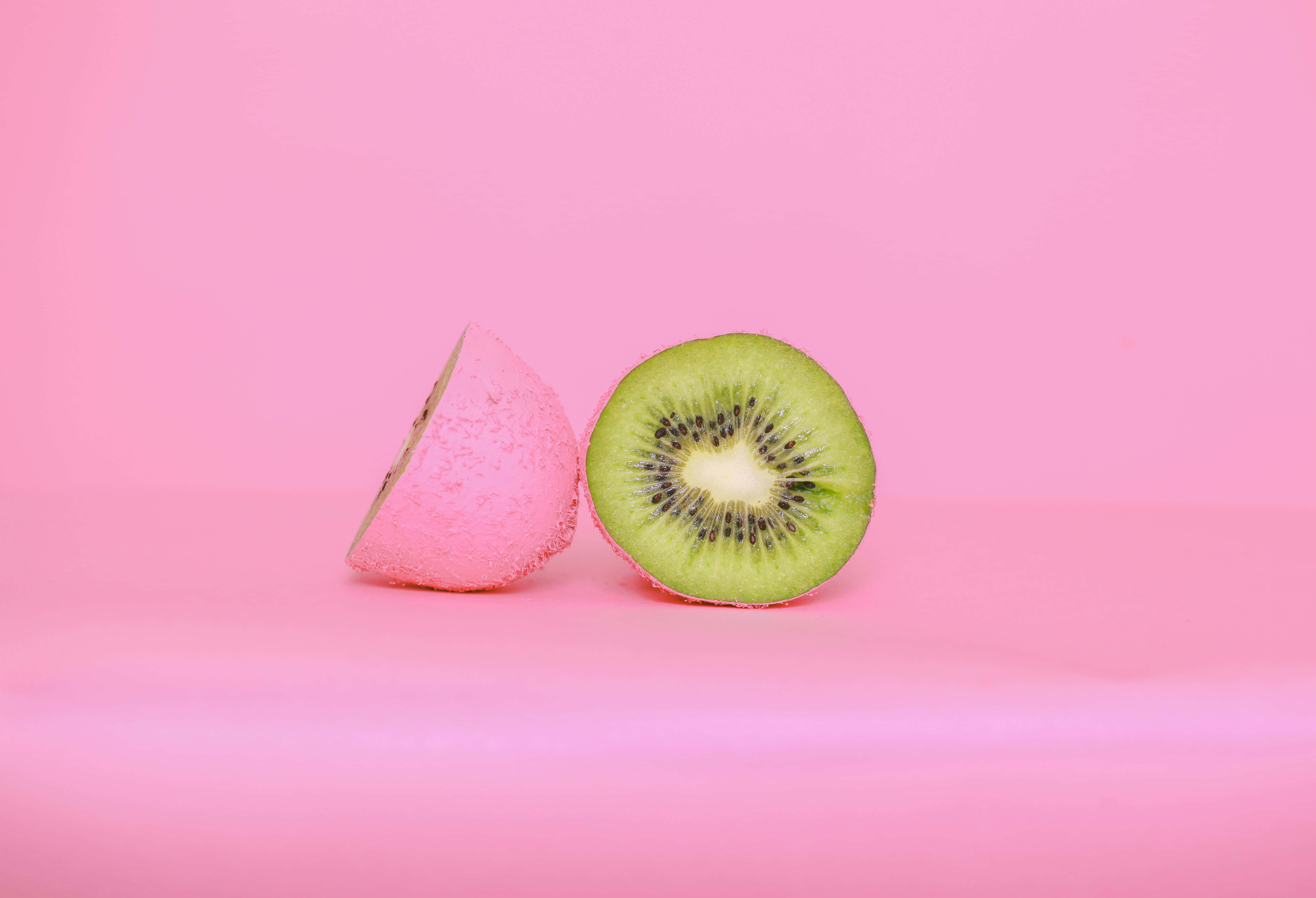 Kiwi 桌布 壁紙 — 一個充滿活力的綠色奇異果切半，露出黑色種子，旁邊放著染成粉紅色的半邊，背景是柔和的粉紅色。