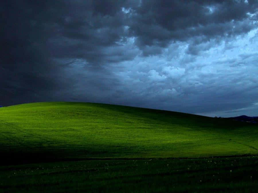 Windows XP 桌布 壁紙 — 一個充滿活力的綠色山丘在戲劇性的暴風雨天空下彎曲，帶有烏雲和藍灰色光線。