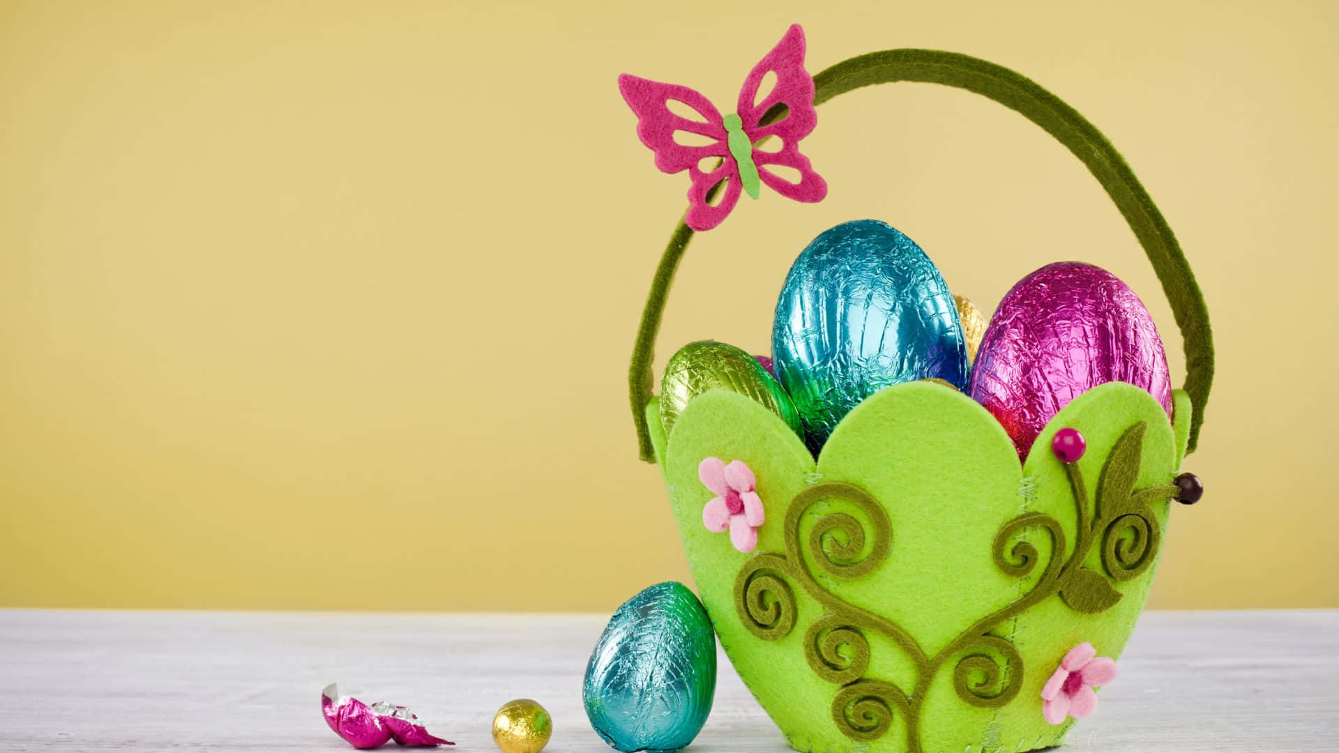 Easter Basket 桌布 壁紙 — 一個充滿活力的綠色氈製復活節籃子，飾有粉紅色花朵和蝴蝶，在柔和的黃色背景上裝著閃亮的箔紙蛋。