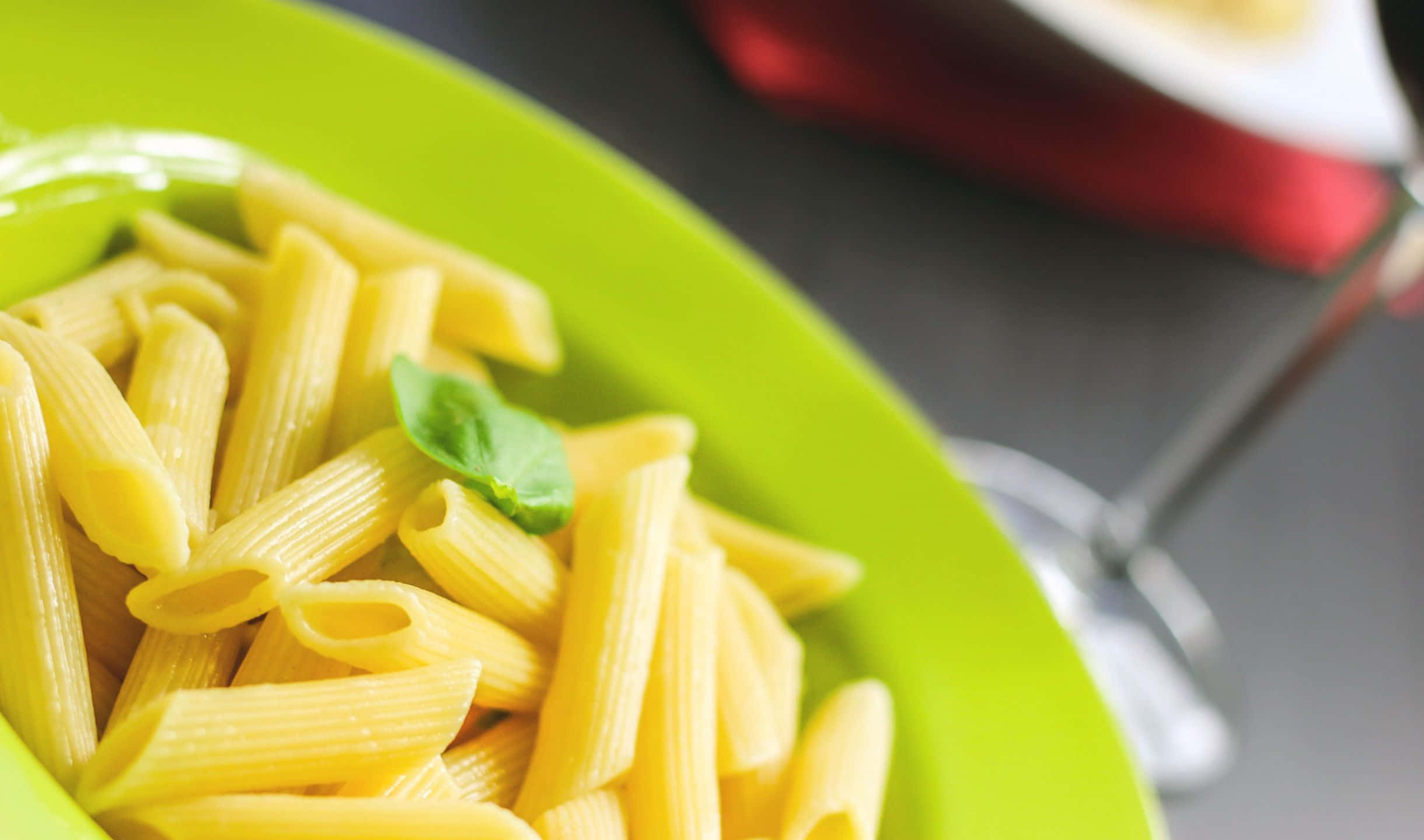 2440x1440 Pasta 桌布 壁紙 — 一個充滿活力的綠色盤子盛裝著金黃色的長通粉，上面放著一片新鮮的羅勒葉。