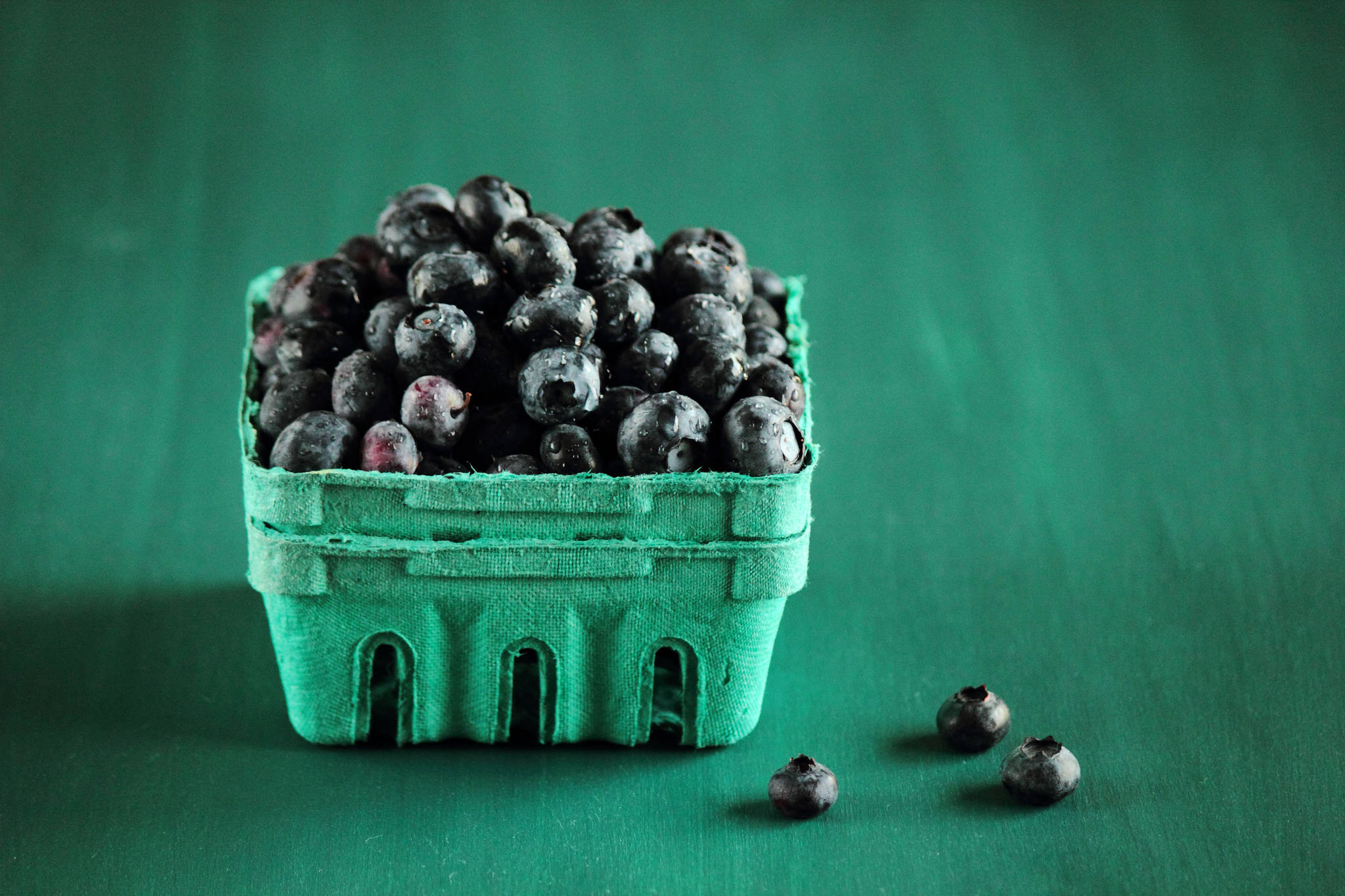 Blueberry 桌布 壁紙 — 一個充滿活力的綠色紙板籃子，裝滿了飽滿的藍莓，放在相配的綠色表面上，附近散落著幾顆漿果。