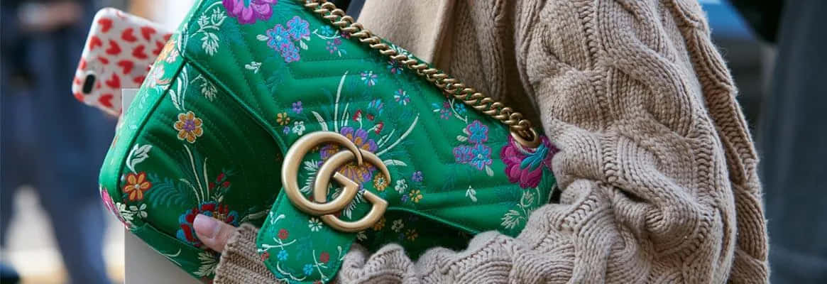 Gucci 桌布 壁紙 — 一個充滿活力的綠色花卉絎縫手提包的特寫，帶有金色五金配件，放在米色電纜針織毛衣上。