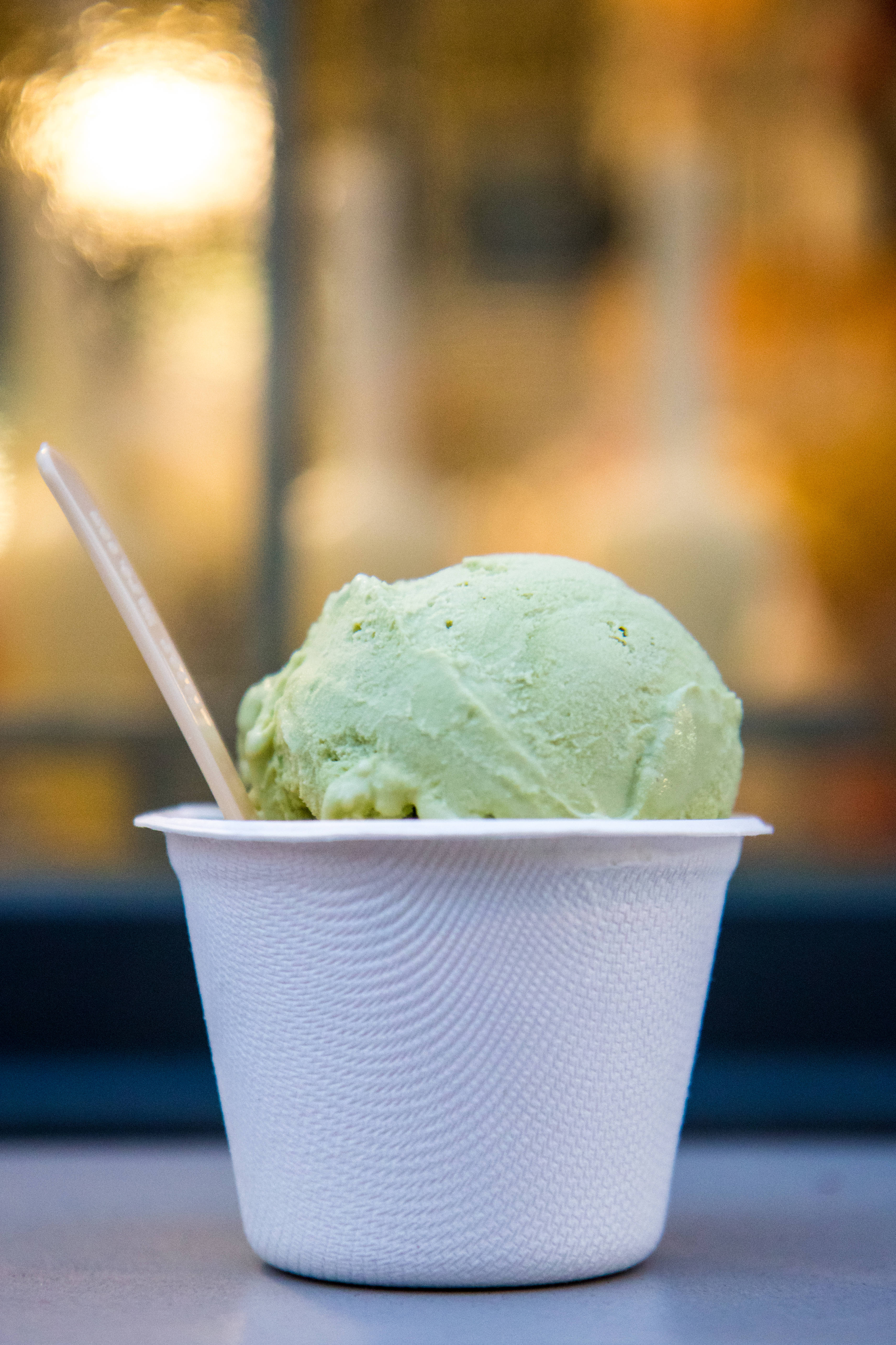 Ice Cream 桌布 壁紙 — 一個充滿活力的綠色開心果義式冰淇淋球放在白色紋理杯中，配以木匙，背景是溫暖的散景。
