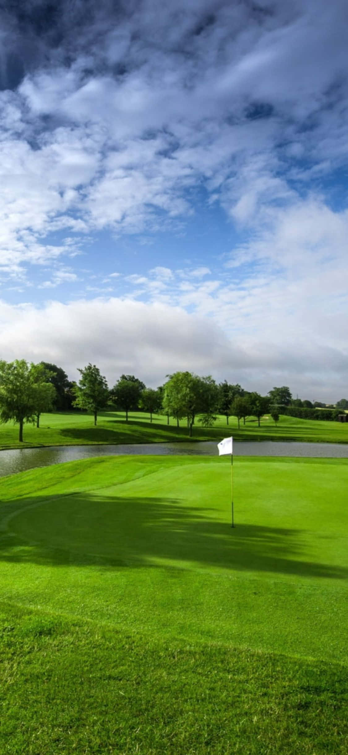 iPhone X Golf Course 桌布 壁紙 — 一個充滿活力的綠色高爾夫果嶺，上面有白色旗幟，在戲劇性的藍天下，充滿蓬鬆的白雲。