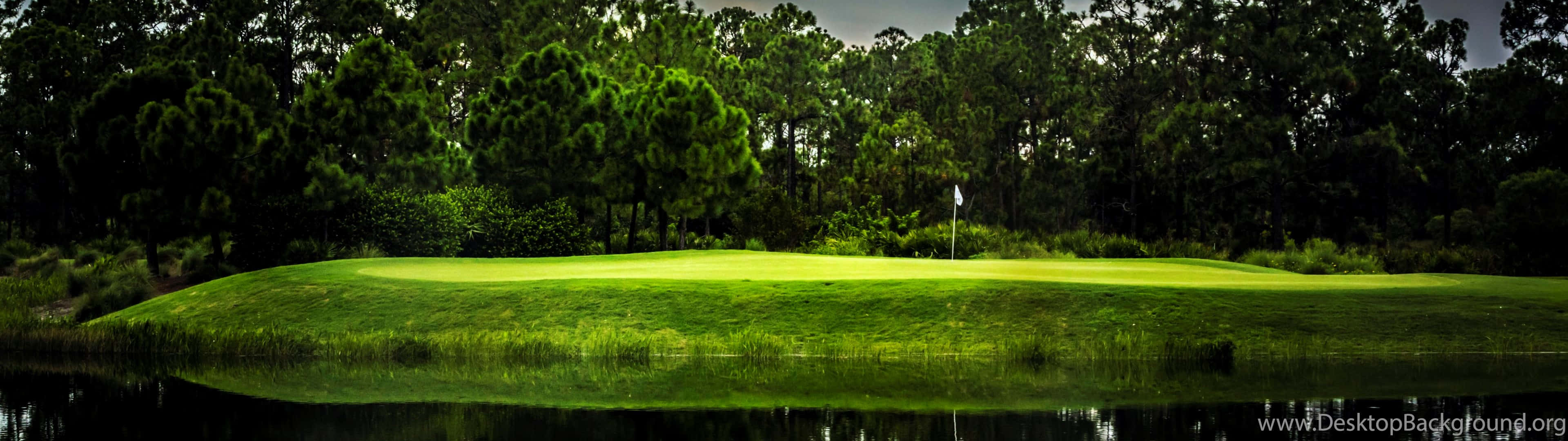 Golf 3840x1080 桌布 壁紙 — 一個充滿活力的綠色高爾夫果嶺，旗桿立在平靜的水障礙旁，天空陰沉。