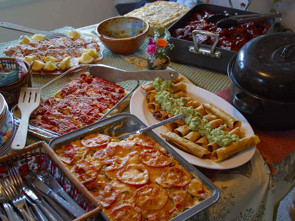 Mexican Food 桌布 壁紙 — 一個充滿活力的自助餐桌，擺滿了起司辣醬玉米餅、塗上酪梨醬的玉米捲餅，以及在溫暖廚房燈光下滋滋作響的肉類。