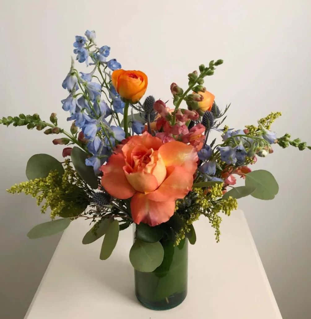 Floral Arrangement 桌布 壁紙 — 一個充滿活力的花卉佈置，包含橙色玫瑰和藍色飛燕草，放在白色桌子上的綠色花瓶裡。