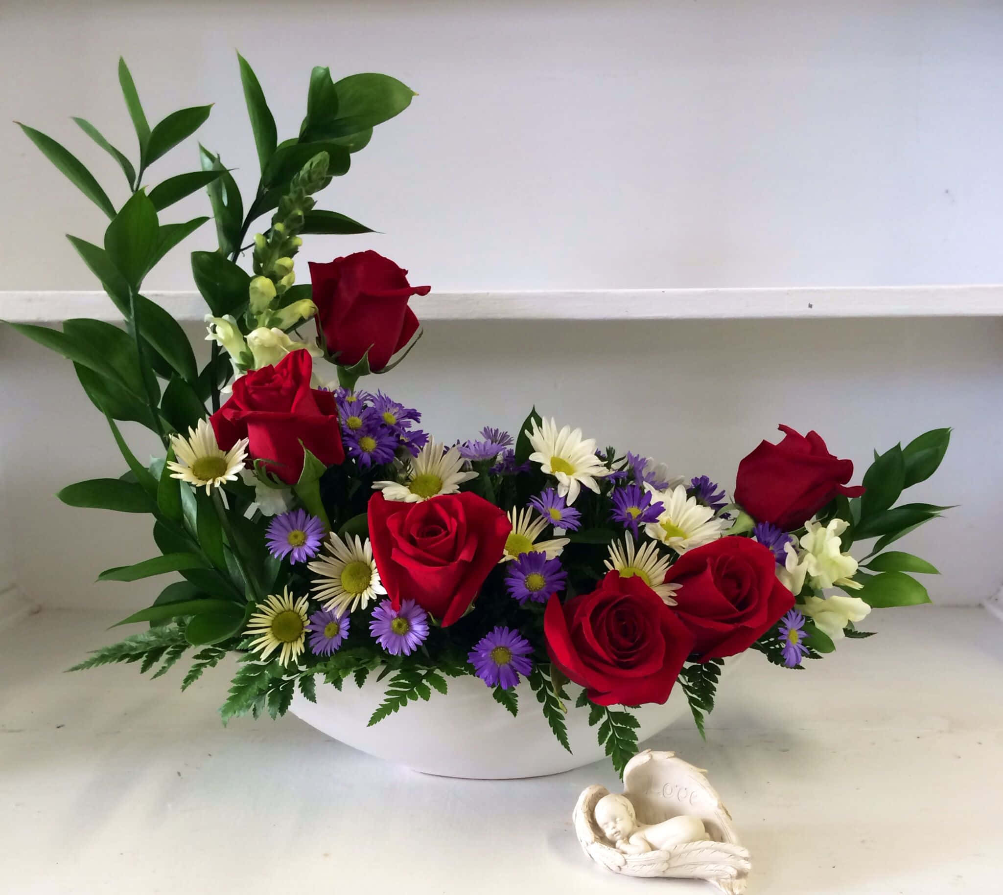 Flower Arrangement 桌布 壁紙 — 一個充滿活力的花卉佈置，包含紅玫瑰、紫色雛菊和綠葉，放在一個白色碗中，旁邊有一個小天使雕像。