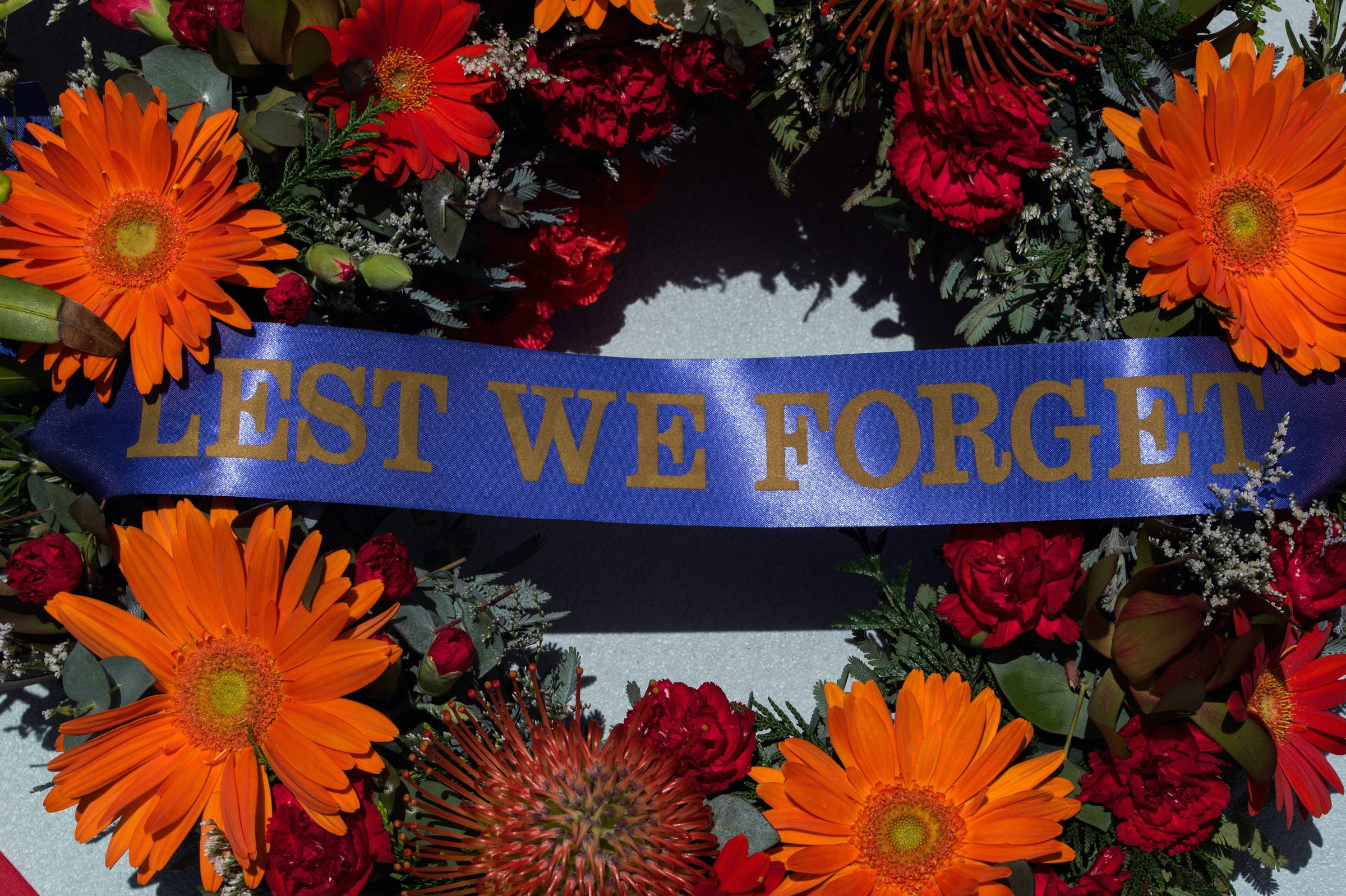 Remembrance 桌布 壁紙 — 一個充滿活力的花圈，由橙色非洲菊和紅玫瑰組成，在灰色表面上拿著一條寫有「LEST WE FORGET」的藍色絲帶。